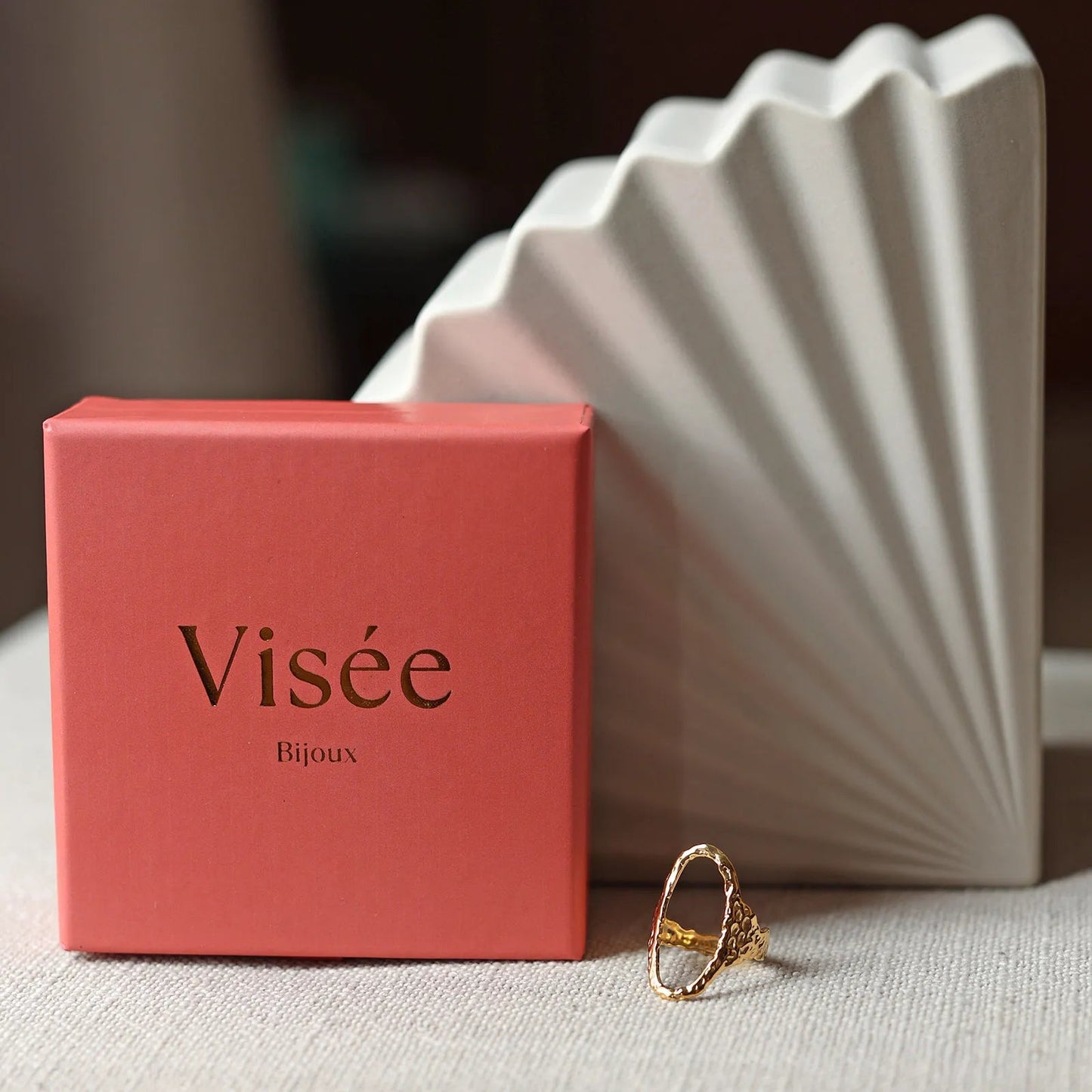Bague Maintenant posée à plat, en acier inoxydable doré, à côté de la boîte Visée Bijoux.