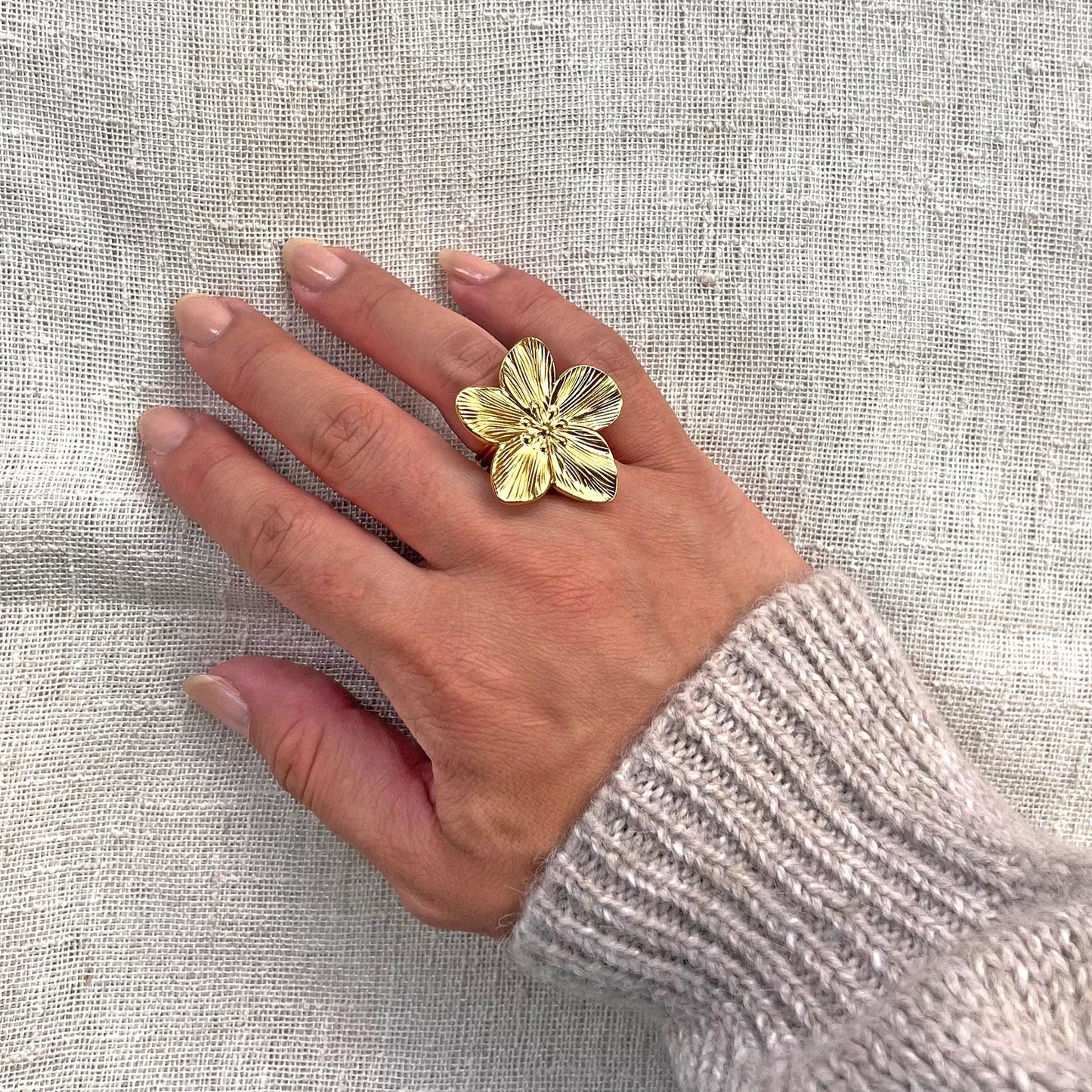 Bague réglable dorée en acier inoxydable, design fleur , portée sur une main féminine avec un pull en maille claire, posée sur un tissu beige texturé.