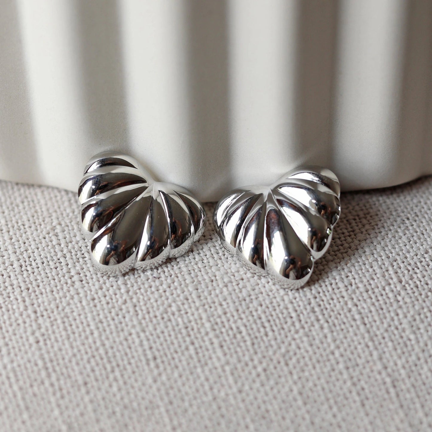 Boucles d'oreilles "Coeur audacieux", en acier inoxydable argenté, au design sculpté en forme de coeur, capturées en gros plan sur fond texturé beige.