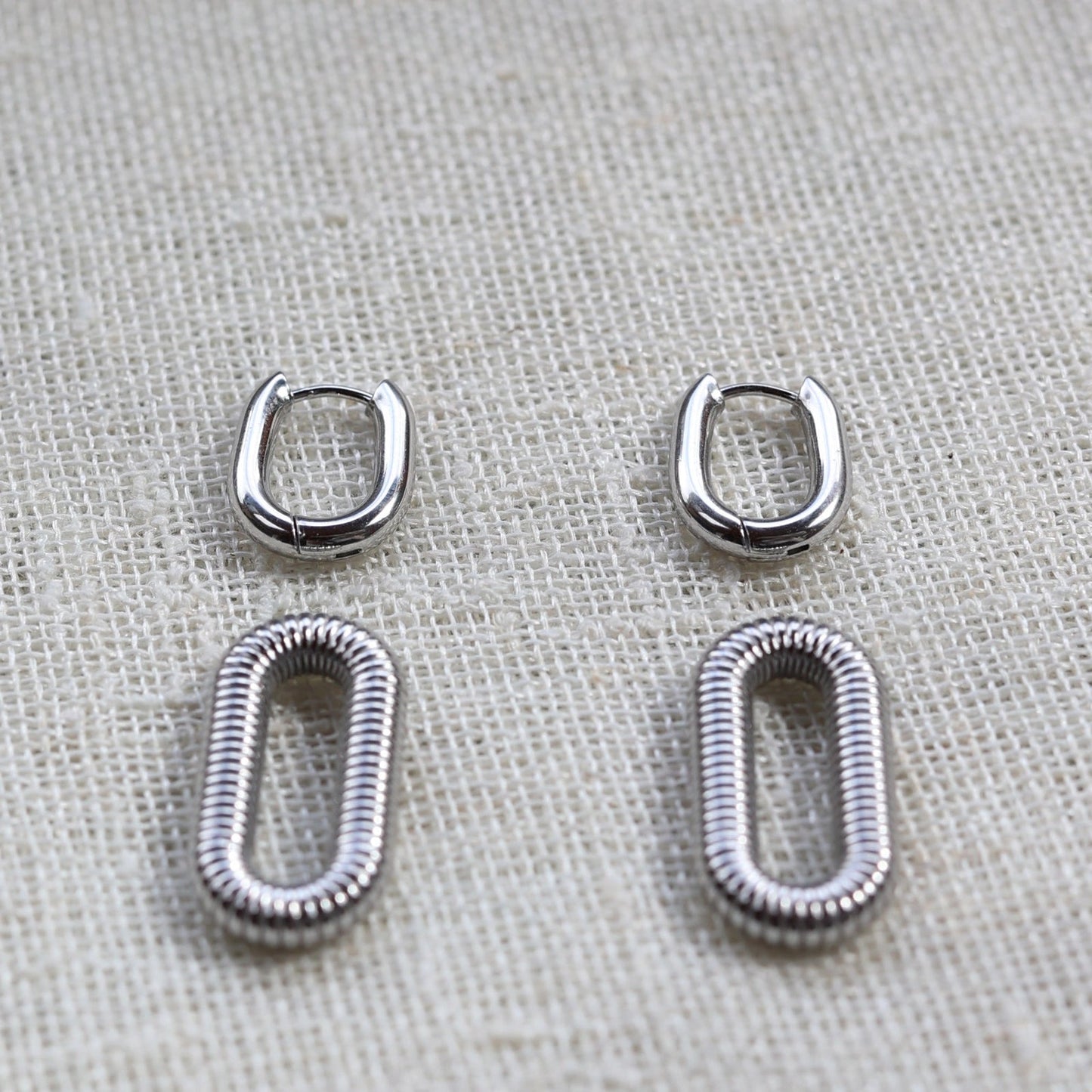 Boucles d'oreilles "J'existe" en acier argenté, au design modulable, photographiées sur un tissu beige texturé.