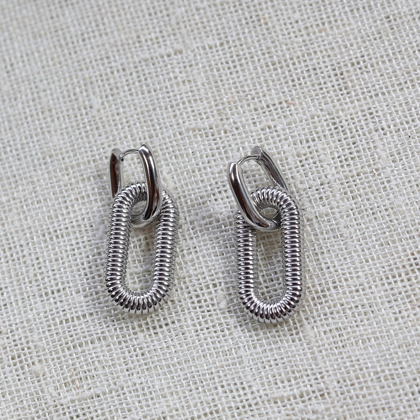 Boucles d'oreilles "J'existe" en acier argenté, au design modulable, photographiées sur un tissu beige texturé.