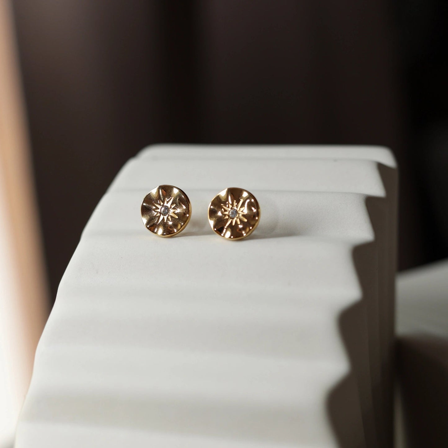 Boucles d’oreilles Céleste dorées motif étoile, serties d'un zircon, posées sur un fond beige.