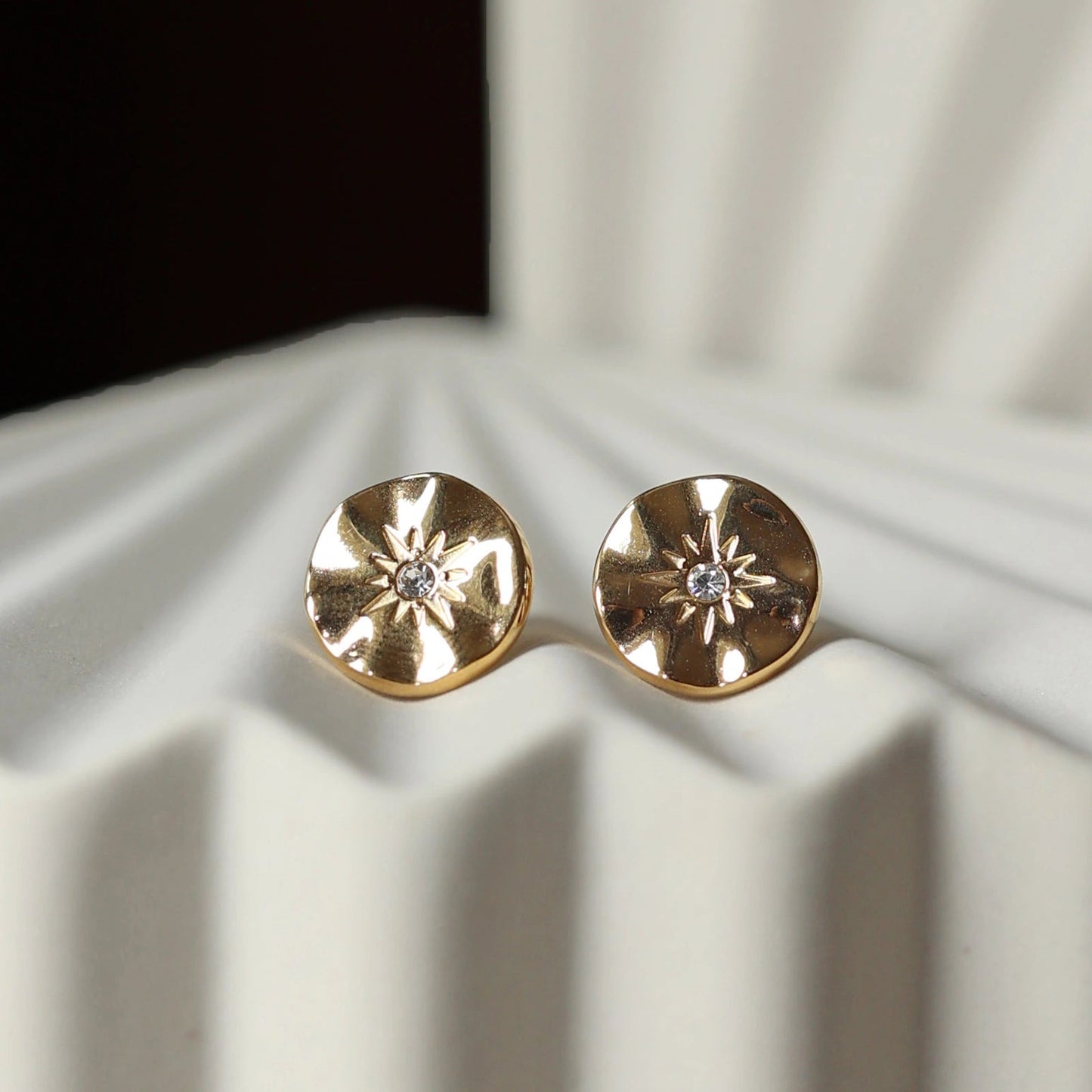 Vue rapprochée des boucles d’oreilles Céleste dorées motif étoile, serties d'un zircon, posées sur un fond beige.