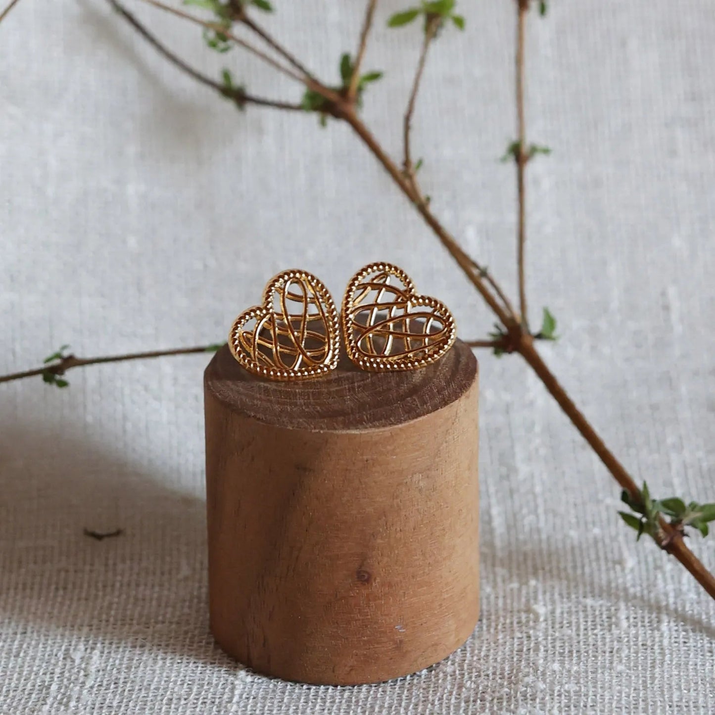 Boucles d'oreilles Etreinte en acier doré, cœurs tressés capturés en gros plan sur un morceau de bois et sur fond de tissu beige texturé et une branche de feuillage