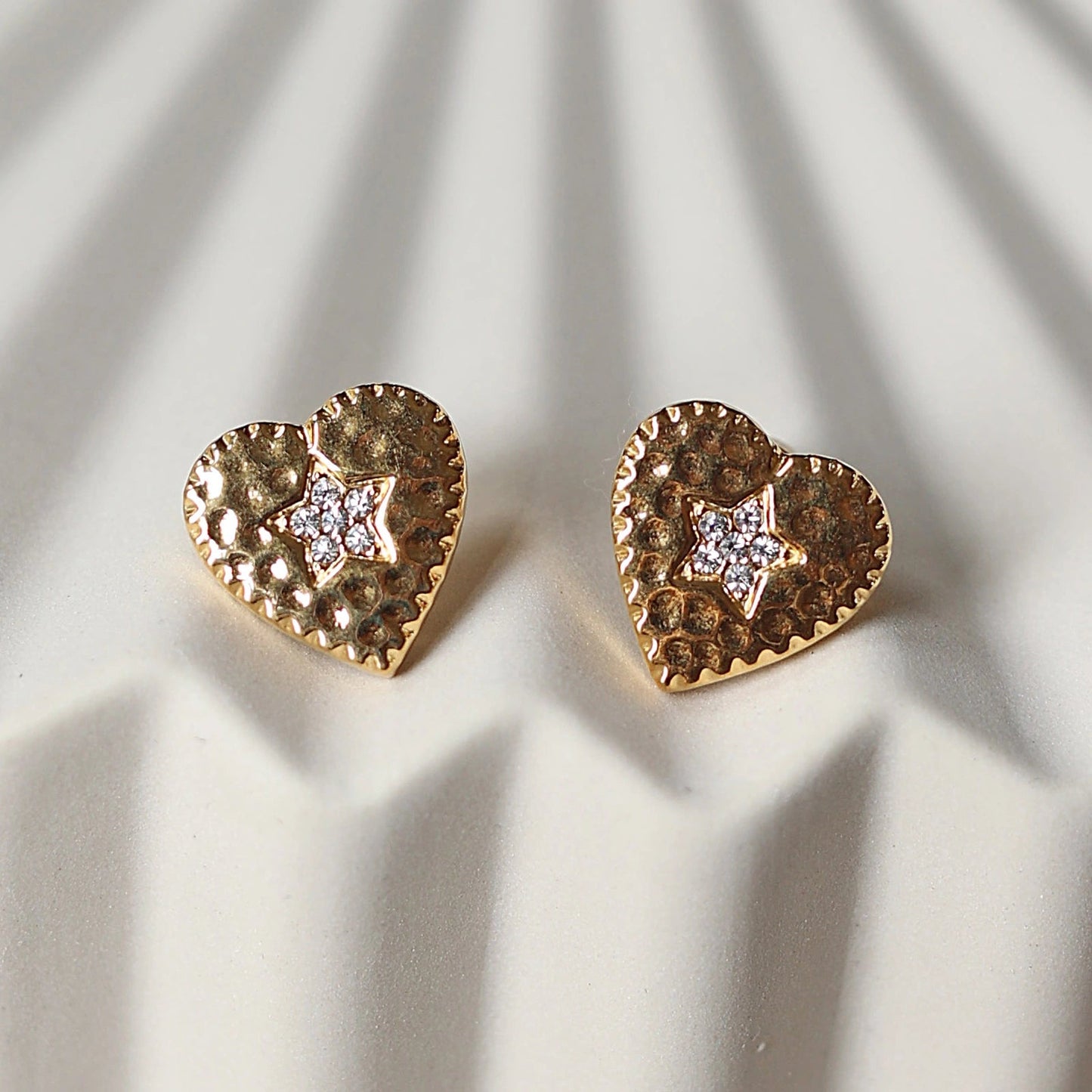 Boucles d'oreilles Promesse, en forme de cœur ornées d'une étoile, en acier inoxydable doré, photographiées en gros plan sur un support beige texturé.
