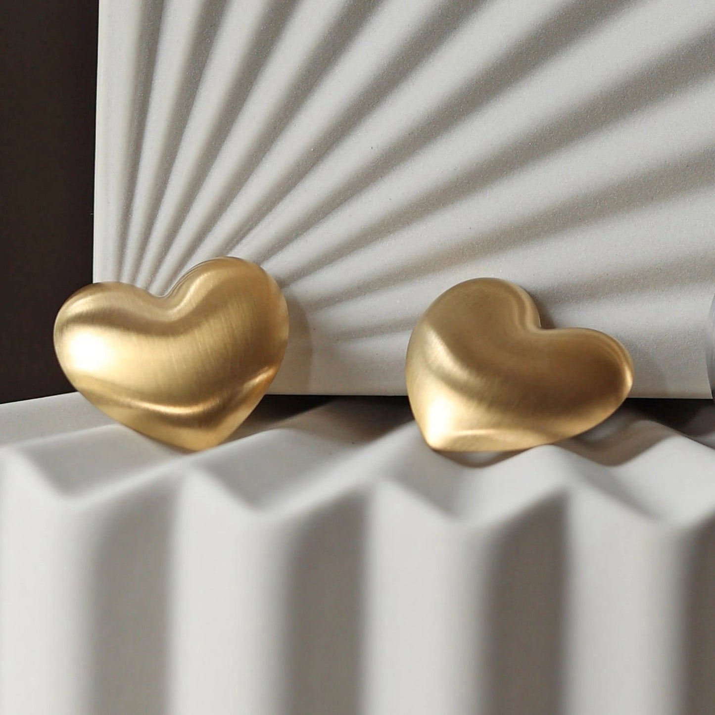 Zoom sur les boucles d'oreilles "S'aimer", en maxi cœurs dorés mat, en acier Inoxydable, posées sur un fond beige texturé.