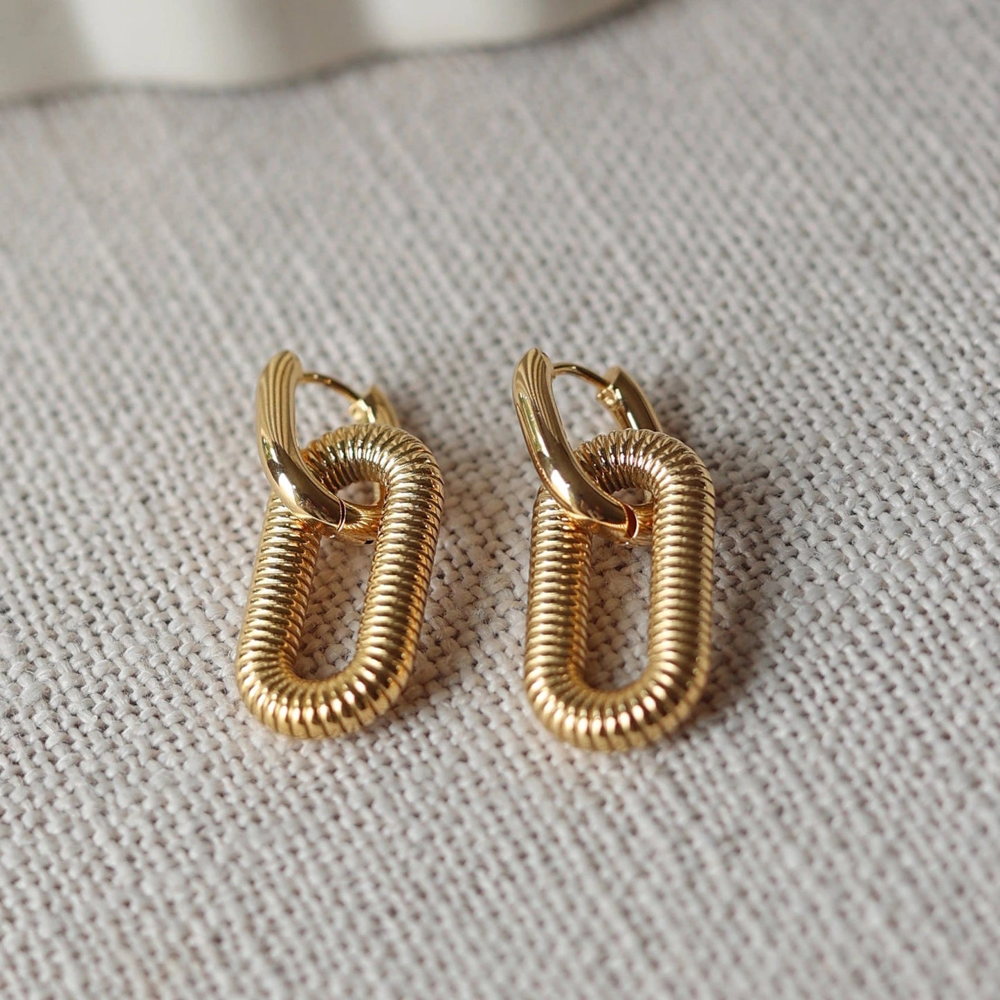 Boucles d'oreilles "J'existe" en acier doré, au design modulable, photographiées sur un tissu beige texturé.
