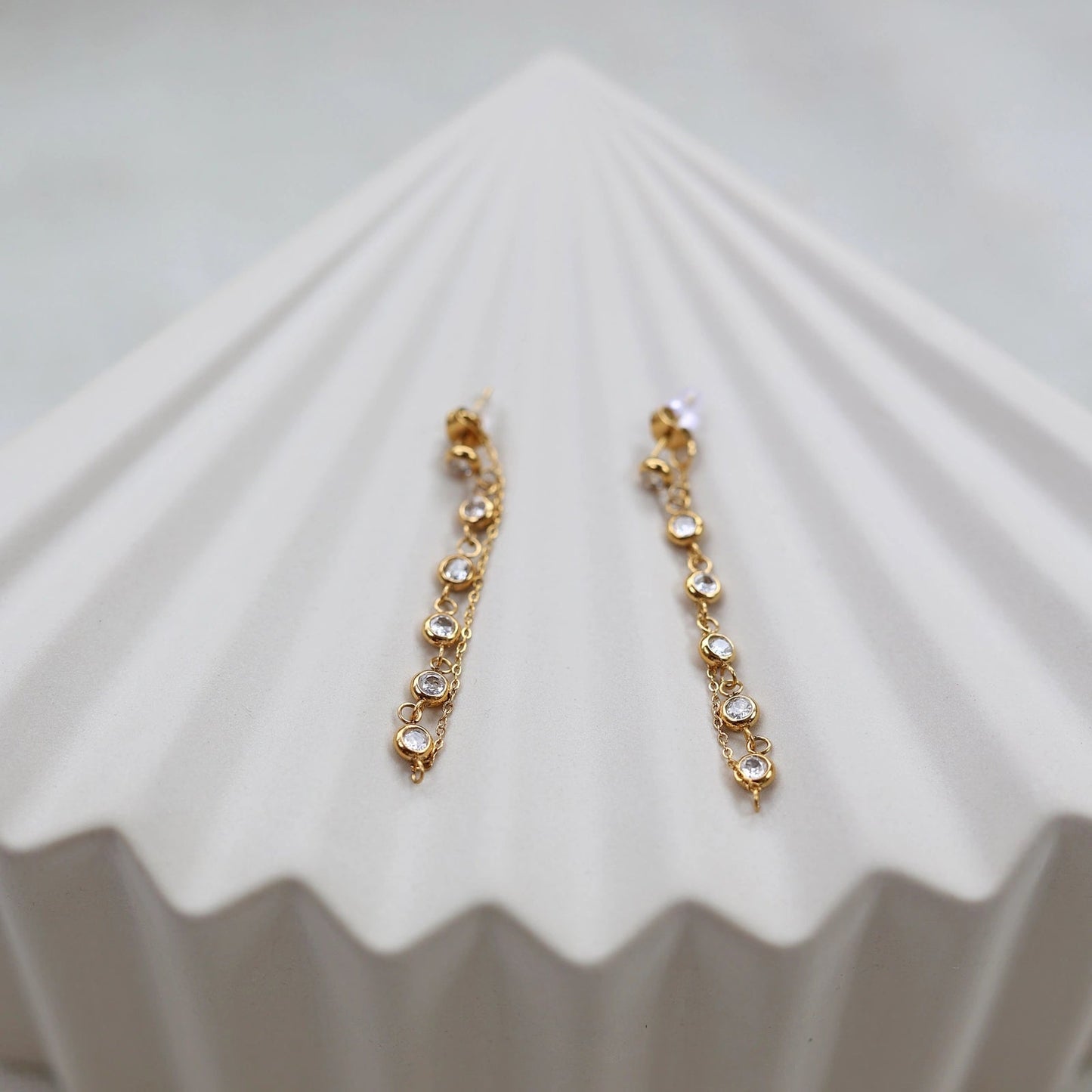 Boucles d'oreilles dorées Eclat, pendantes, serties de strass brillants, en acier. Photographiées sur un support beige texturé.