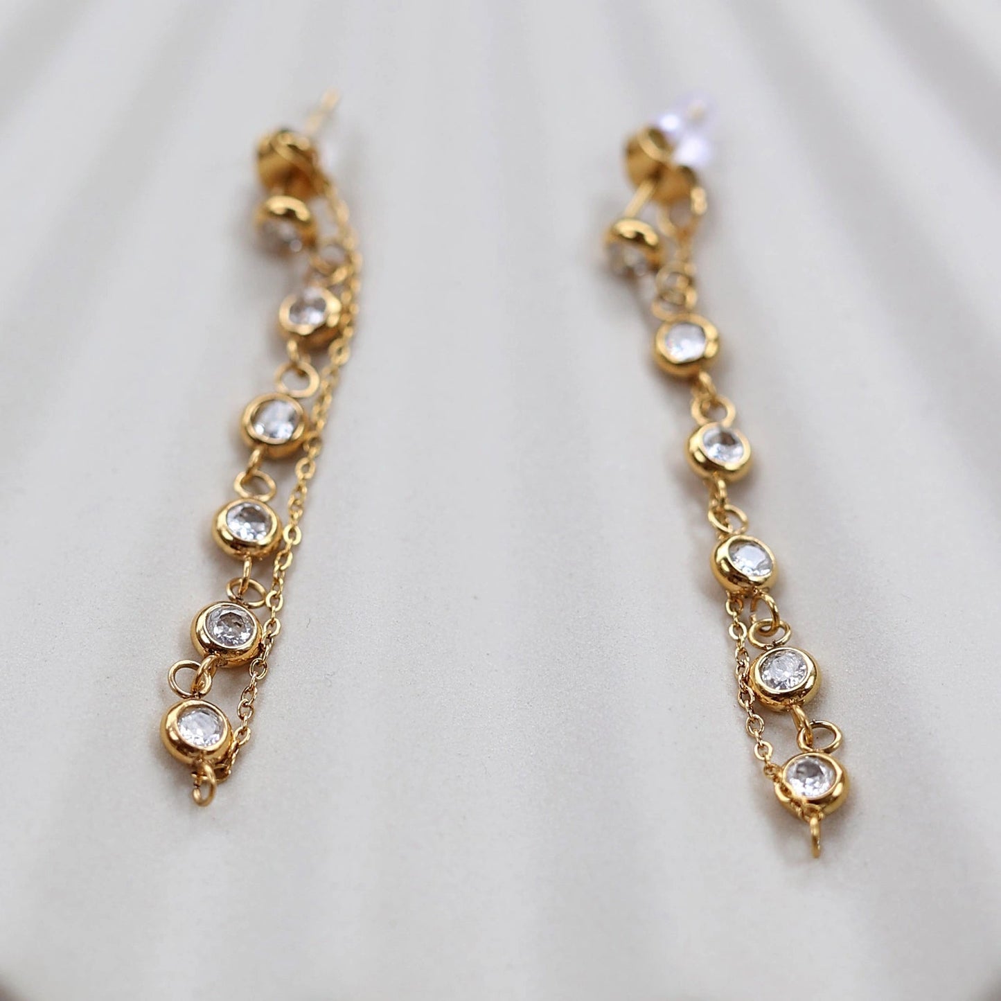 Boucles d'oreilles dorées Eclat, pendantes et serties de strass brillants, en acier. Photographiées en gros plan sur un support beige texturé.