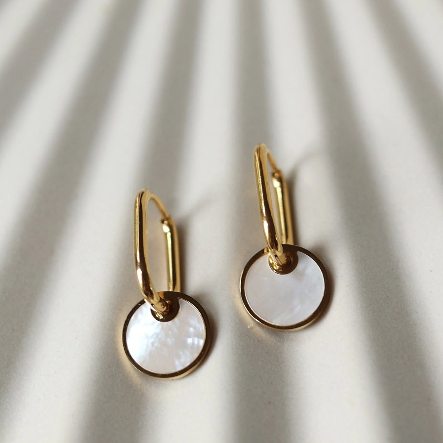 Zoom sur les boucles d'oreilles "Solstice", en acier doré, pendentifs cercles personnalisables à effet nacré, posées sur un fond beige texturé.