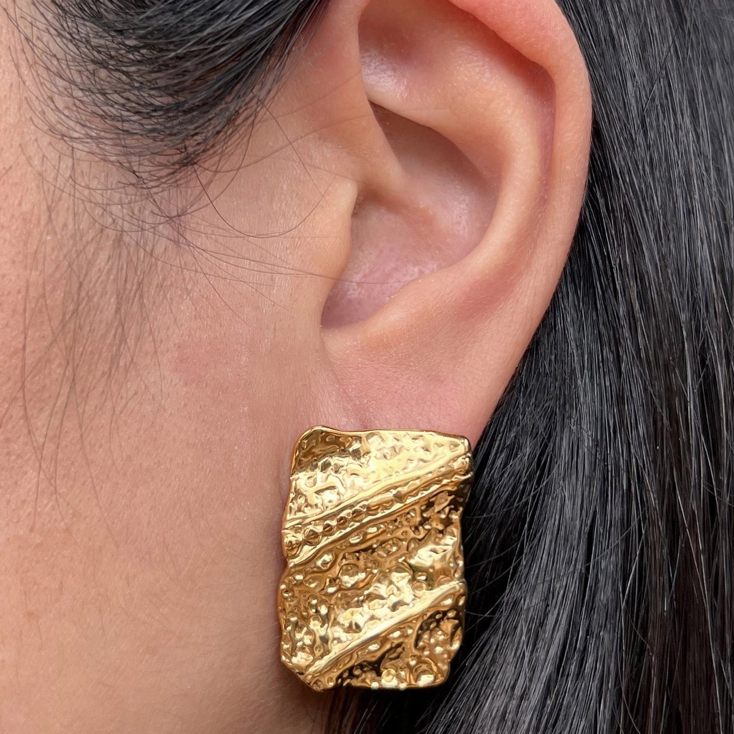 Boucles d'oreilles Affirmation en acier doré, aux formes rectangulaires texturées, portées à l'oreille.
