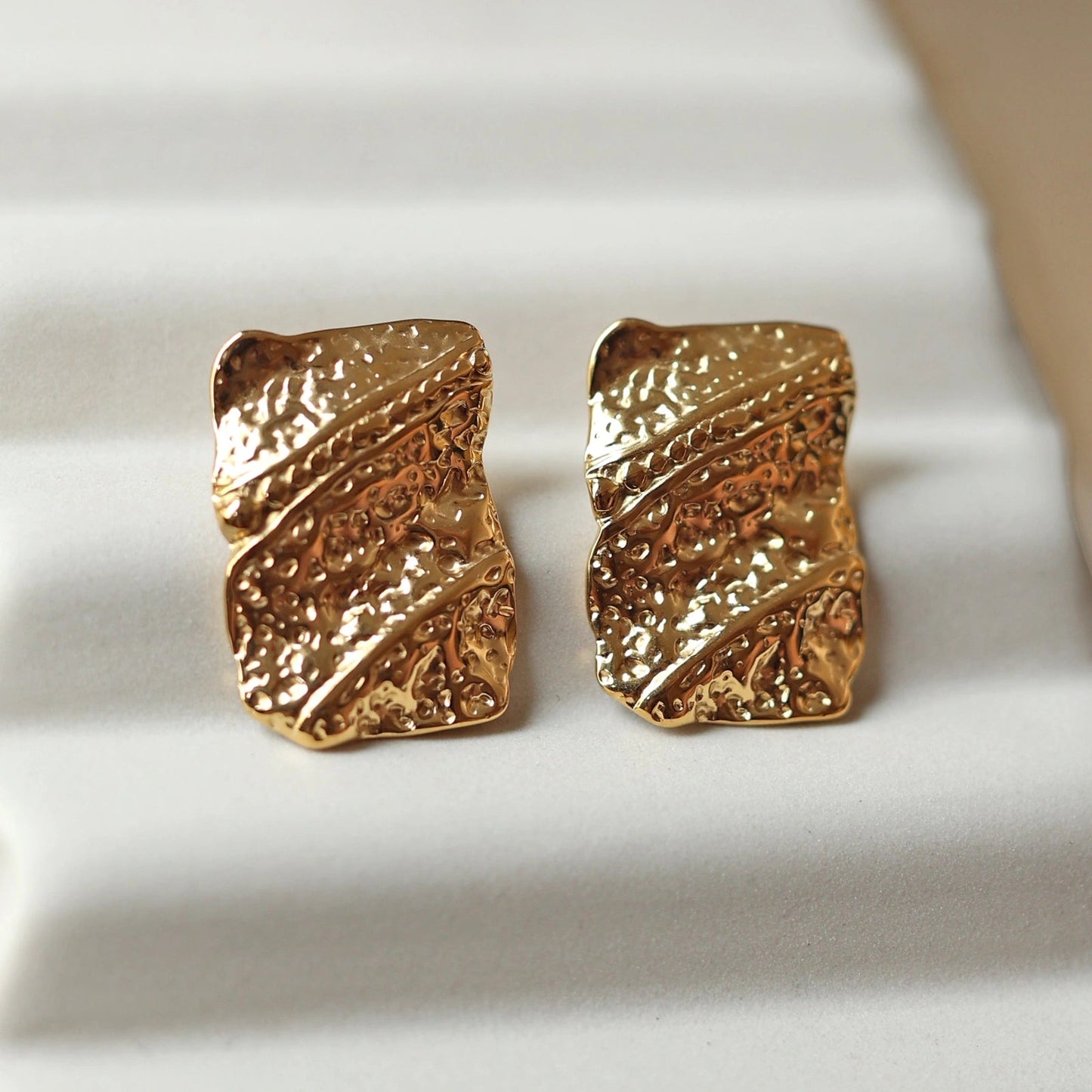 Zoom sur les boucles d'oreilles Affirmation, rectangulaires et texturées en acier doré, posées sur un fond beige texturé.
