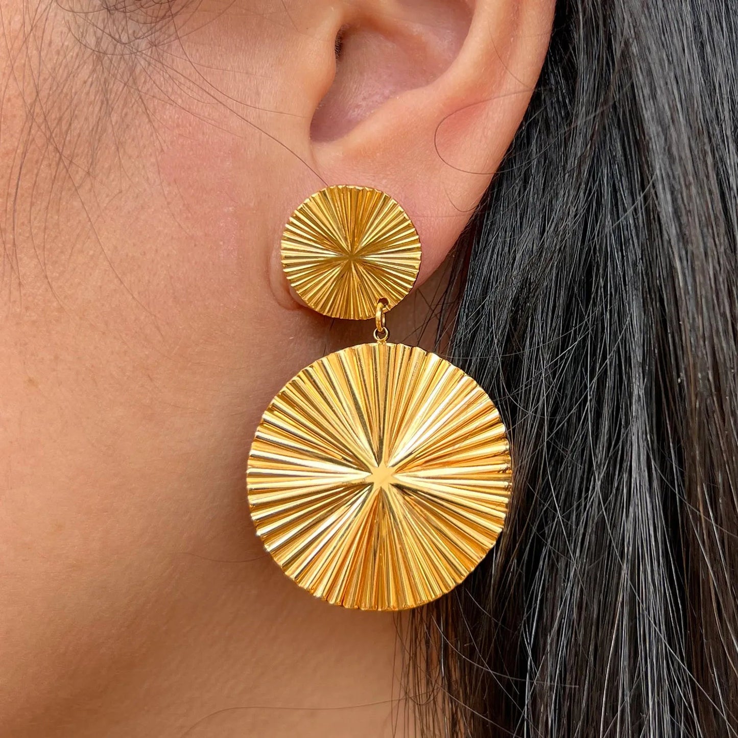 Boucles d'oreilles Vivante pendantes au design solaire strié en acier inoxydable doré, portées sur un modèle, signé Visée Bijoux.