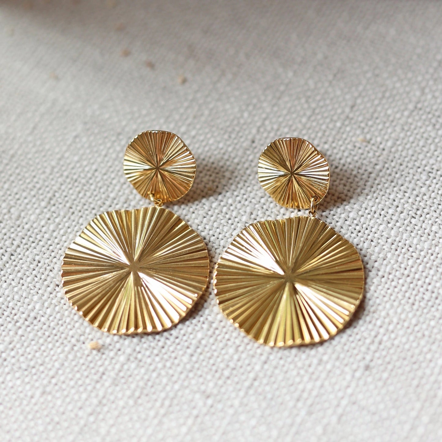 Vue rapprochée des boucles d'oreilles Vivante pendantes au design solaire strié en acier inoxydable doré, posées à plat sur un fond beige texturé.