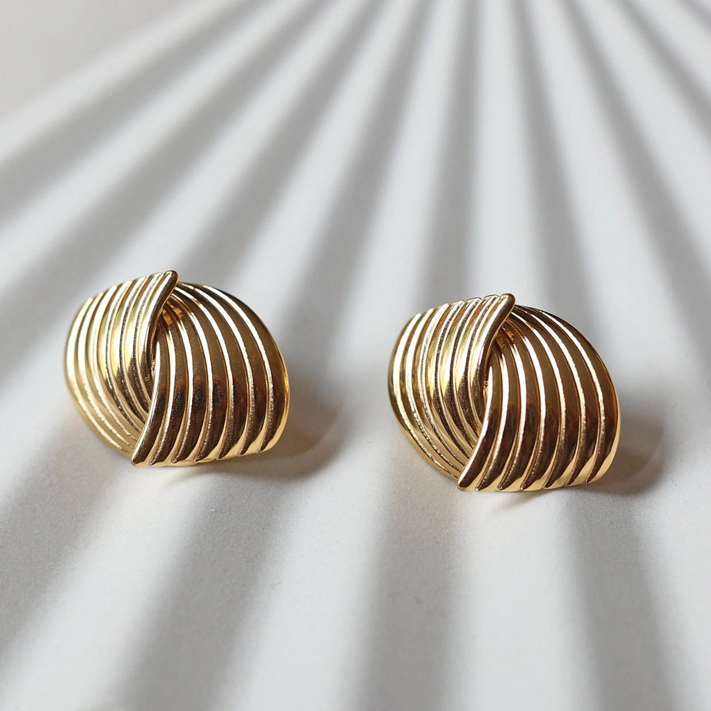 Vue rapprochée des boucles d’oreilles Metémorphose torsadées en acier inoxydable doré, posées à plat sur une structure beige texturée.