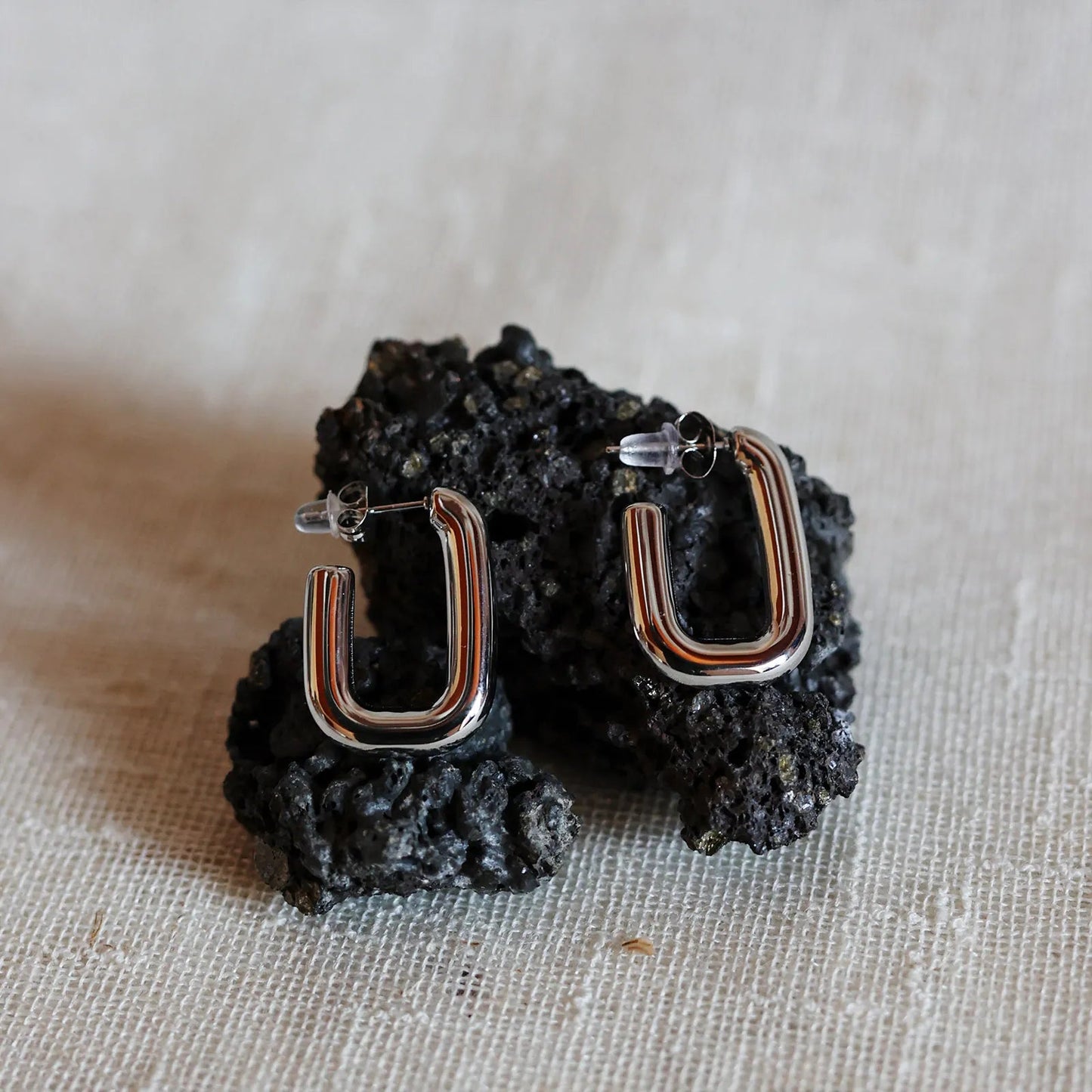 Boucles d'oreilles J'agis, créoles rectangulaires en acier inoxydable argenté, photographiée en vue rapprochée, posées sur une pierre volcanique noire.