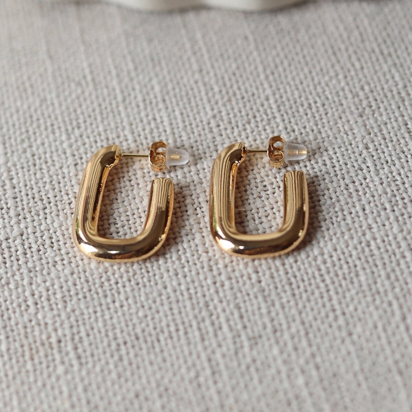 Boucles d'oreilles J'agis, créoles rectangulaires en acier inoxydable doré, photographiée en vue rapprochée, posées sur une toile beige texturée.