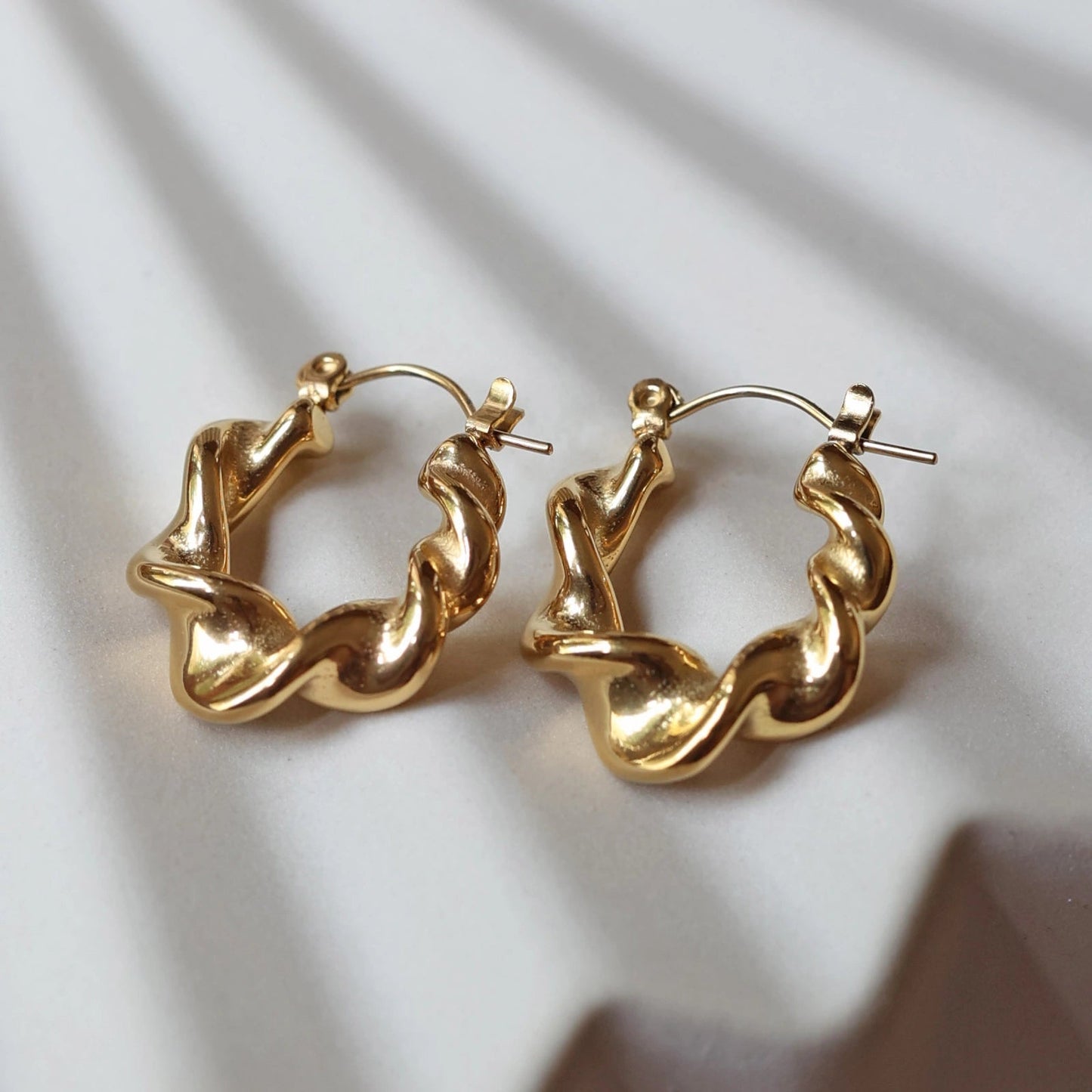 Zoom sur les boucles d’oreilles Elégante - créoles dorées ondulées en acier inoxydable, posées sur un support beige texturé.