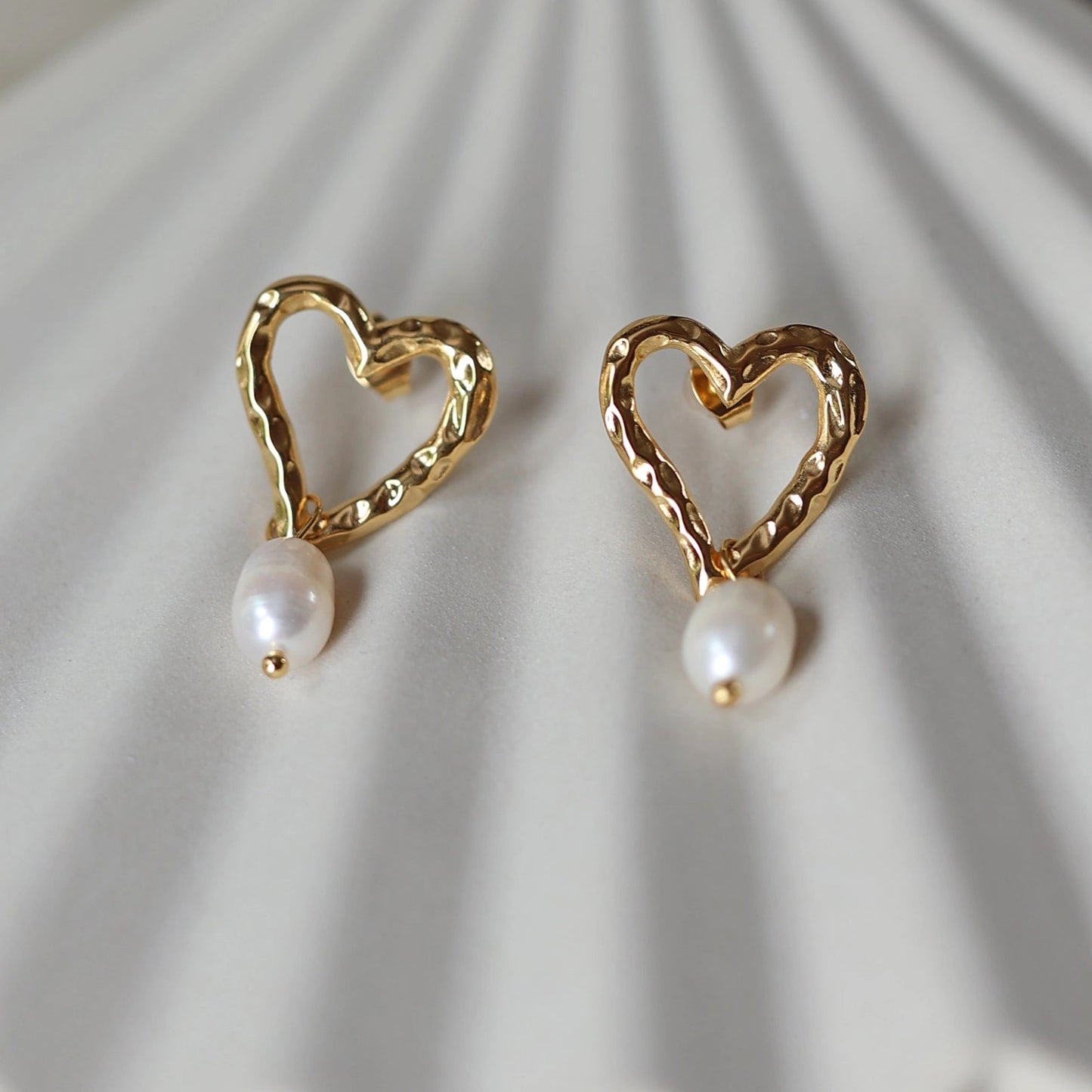 Boucles d’oreilles Loyale, en forme de cœur doré avec perle blanche, en acier inoxydable, photographiées de face sur un fond beige texturé.