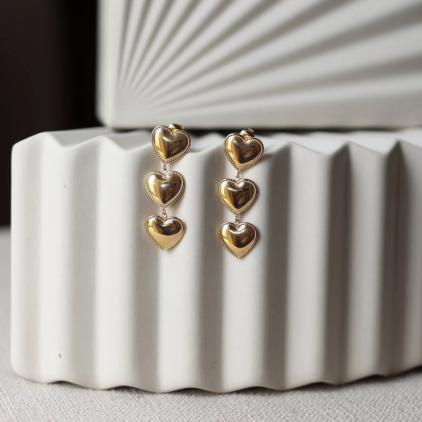 Boucles d'oreilles "Unique", en acier inoxydable doré, pendantes en coeurs, photographiées sur un fond beige texturé.