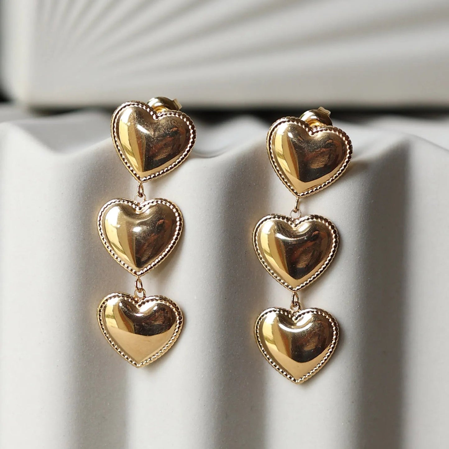 Boucles d'oreilles "Unique", en acier inoxydable doré, pendantes en coeurs, photographiées sur un fond beige texturé.