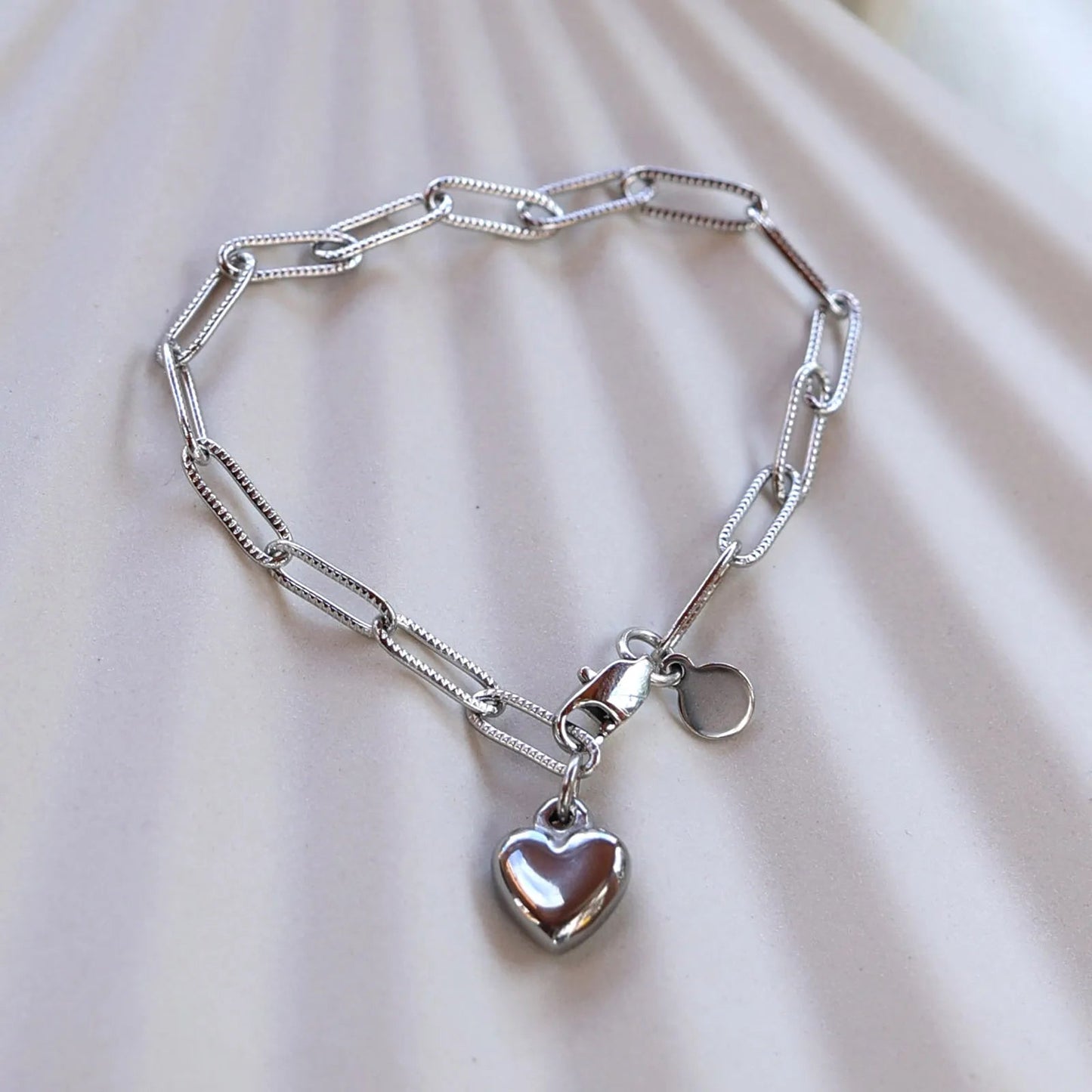 Bracelet Valeur acier inoxydable argenté, avec breloque cœur personnalisable, posé sur un fond beige texturé.