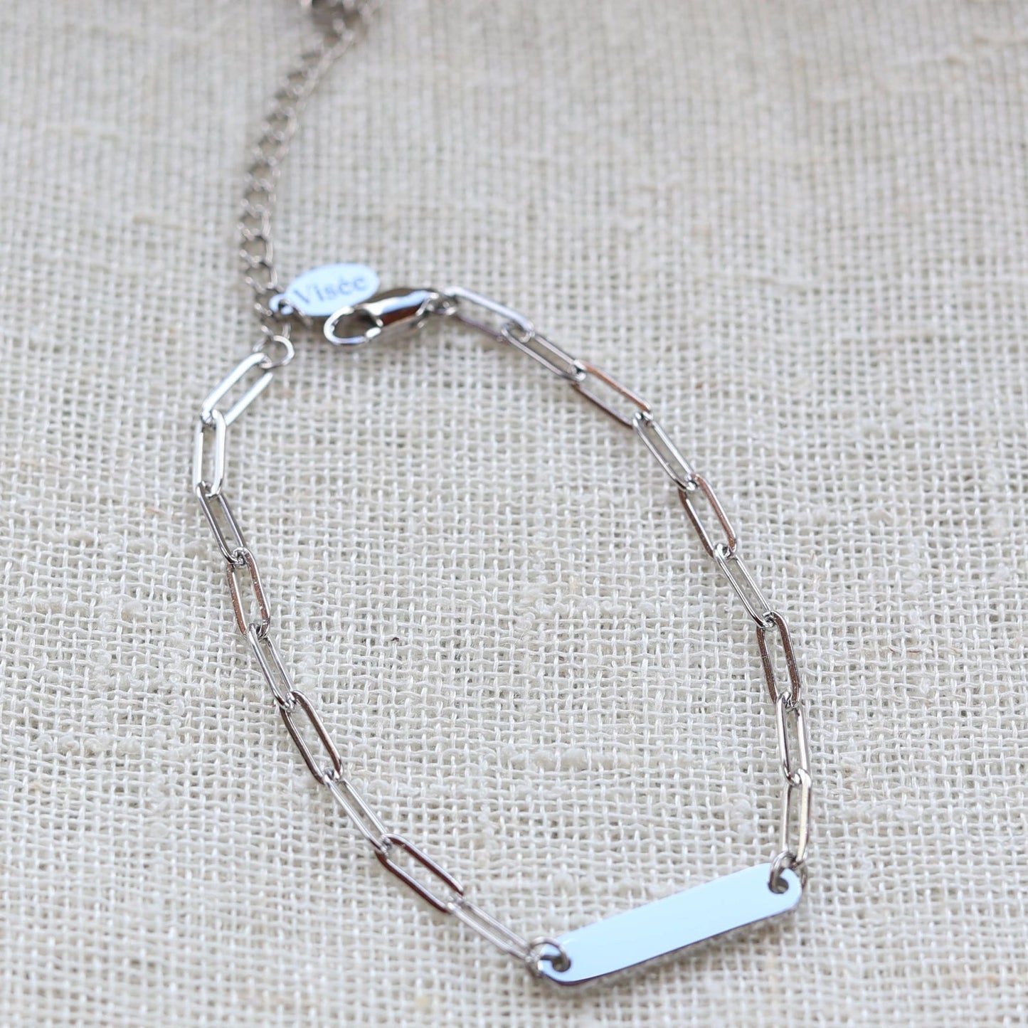 Bracelet "Expression" en acier inoxydable argenté, à personnaliser avec une plaque gravable, présenté sur un fond beige texturé.