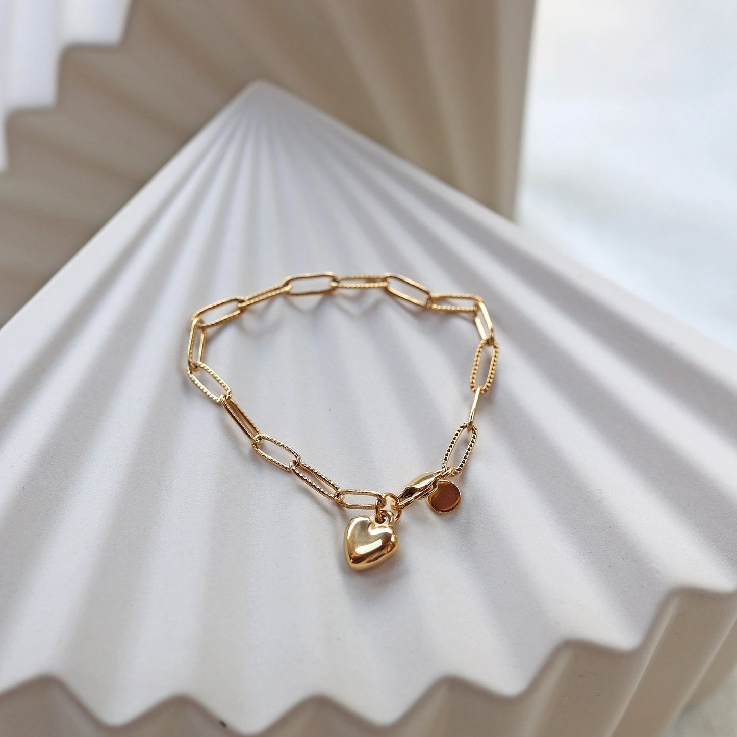 Bracelet Valeur acier inoxydable doré, avec breloque cœur personnalisable, posé sur un fond beige texturé.