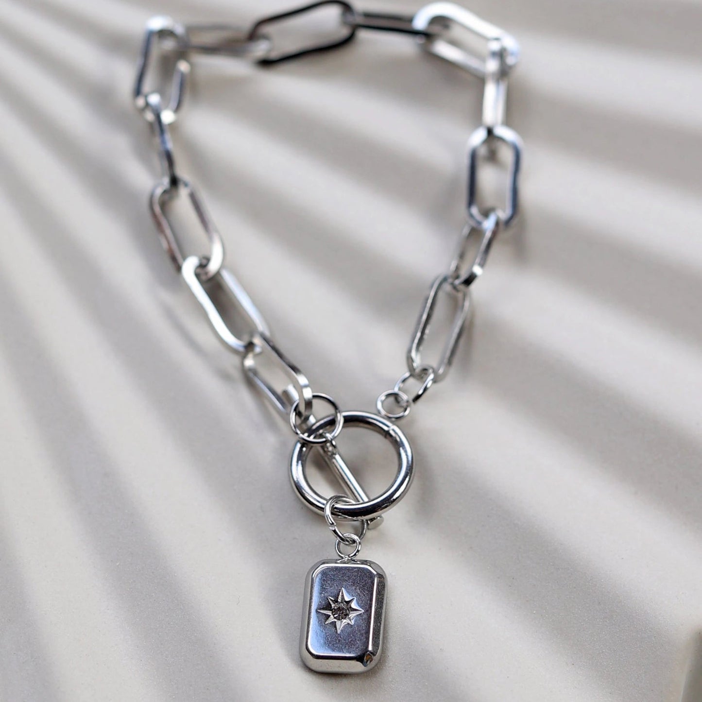 Zoom sur le bracelet "Révélation" en acier argenté, orné d'une breloque rectangulaire personnalisable, posé sur un fond beige texturé.