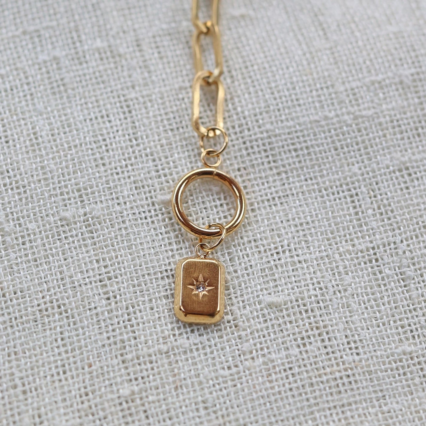 Zoom sur la breloque rectangulaire personnalisable en acier doré, posé sur un fond beige texturé.