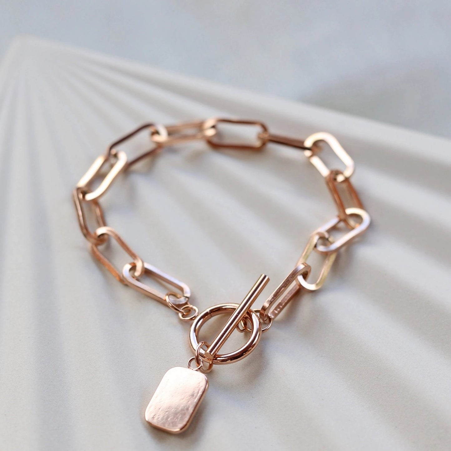 Bracelet "Révélation" en acier rosé avec breloque rectangulaire personnalisable, posé sur un fond beige texturé.