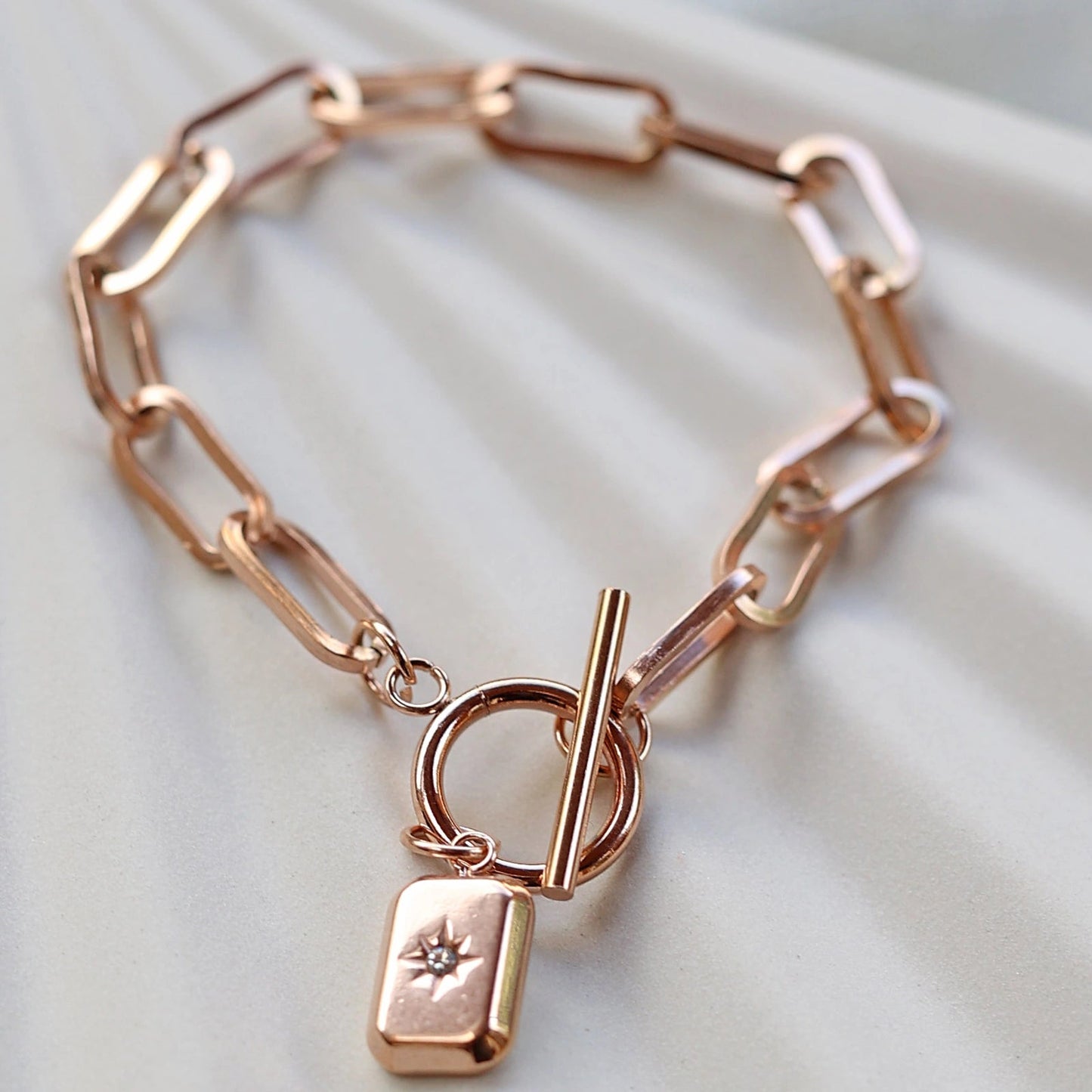 Zoom sur le bracelet "Révélation" en acier rosé avec breloque rectangulaire personnalisable, posé sur un fond beige texturé.