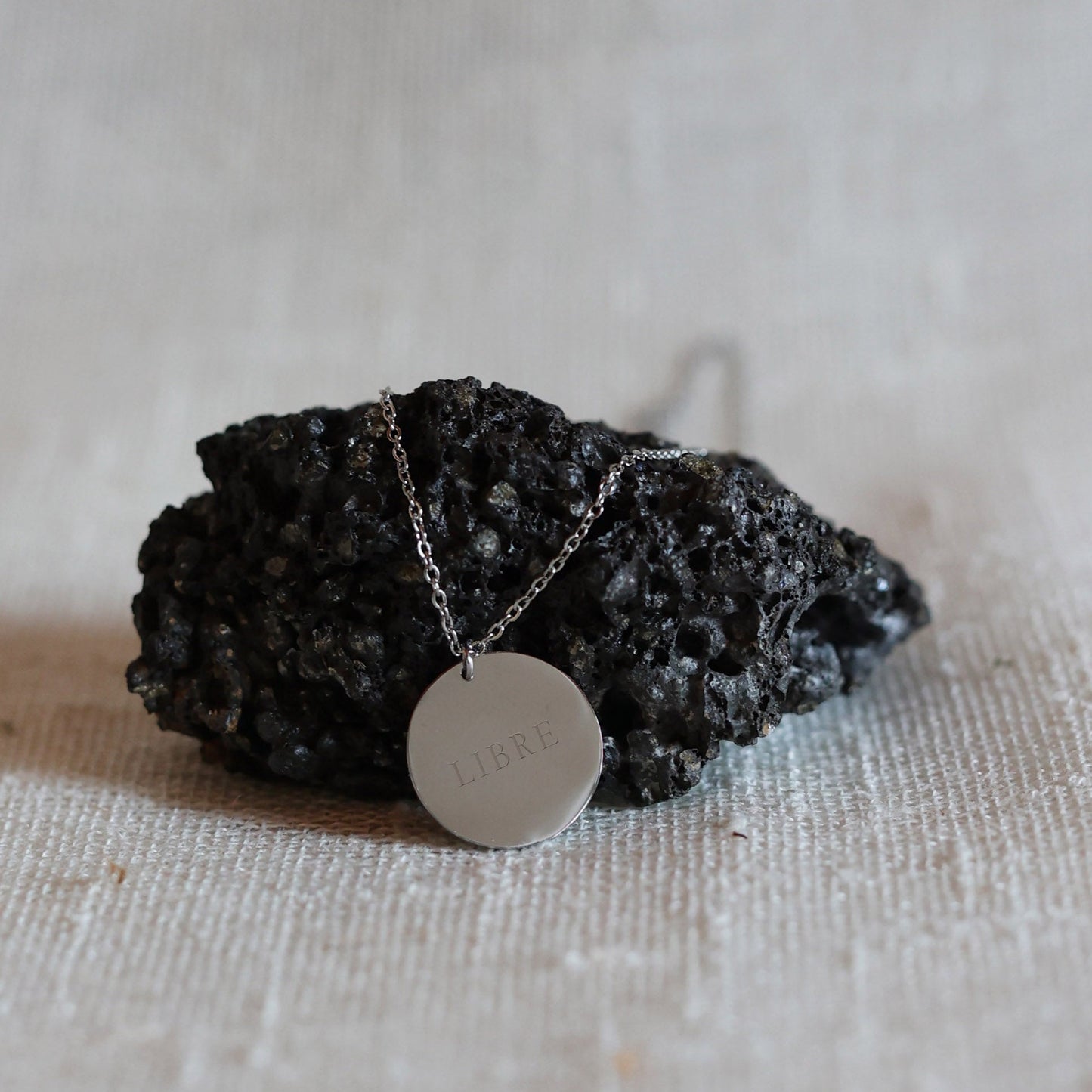Collier "Reflet" avec médaille ronde personnalisé en acier inoxydable argenté, personnalisé LIBRE, posé sur une pierre noire volcanique
