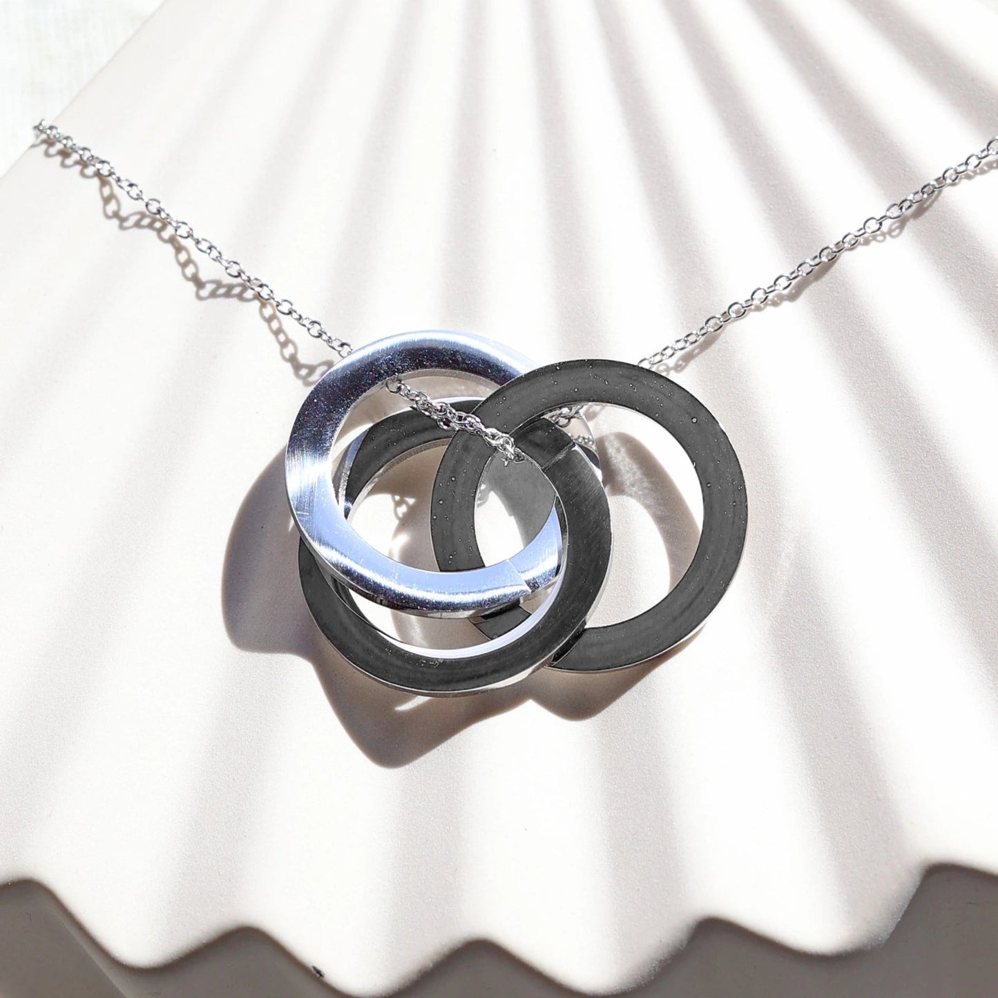 Zoom sur le collier "Je deviens" en acier argenté, avec trois cercles entrelacés personnalisables, posé sur un fond beige texturé.