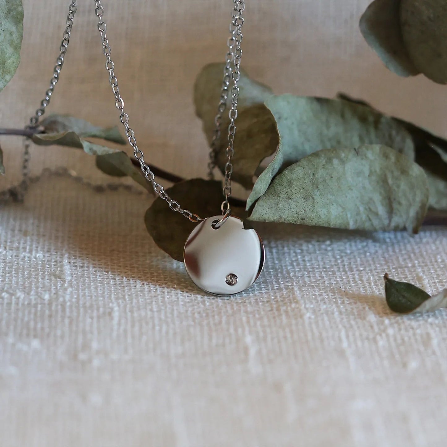 Collier personnalisable "Lumineuse", orné d'une médaille ronde brillante en acier argenté, posé sur un fond beige texturé.