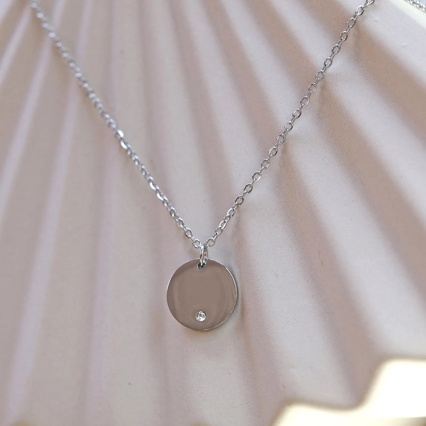 Zoom sur le collier personnalisable "Lumineuse" avec médaille ronde brillante en acier argenté, posé sur un fond beige texturé.