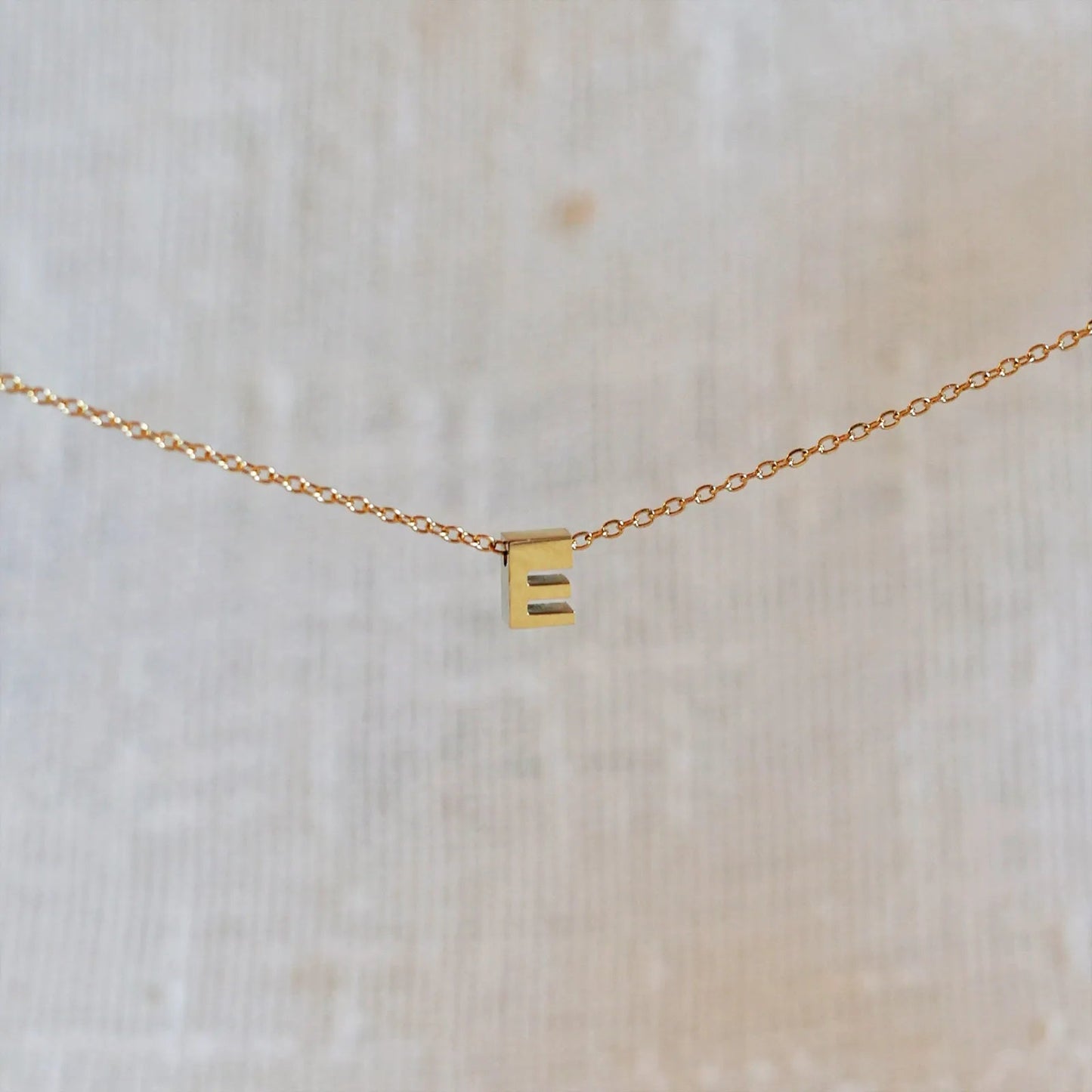 Collier L'instant avec pendentif lettre E doré en acier inoxydable, suspendu au centre d'une maille fine sur fond beige.