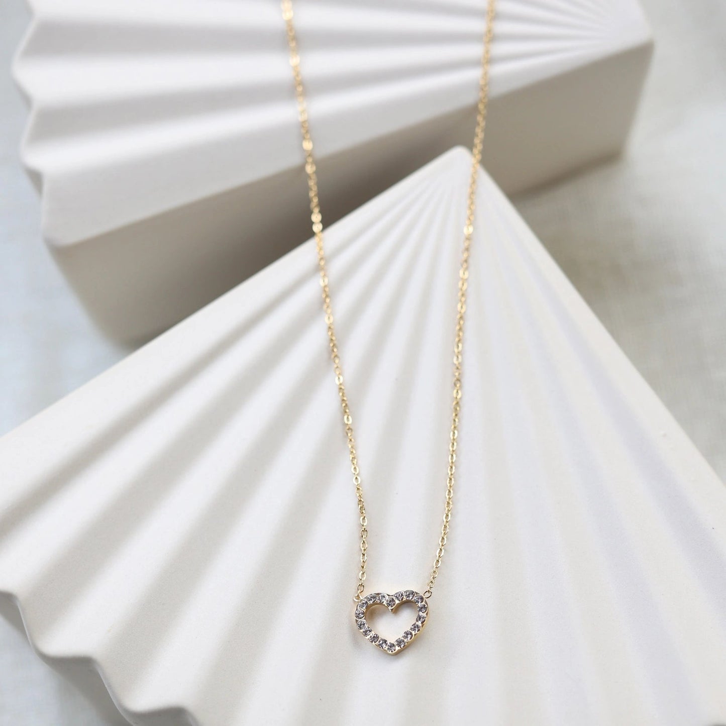 Collier Sentiment, pendentif cœur serti de brillants en acier inoxydable doré, posé sur un support beige texturé.