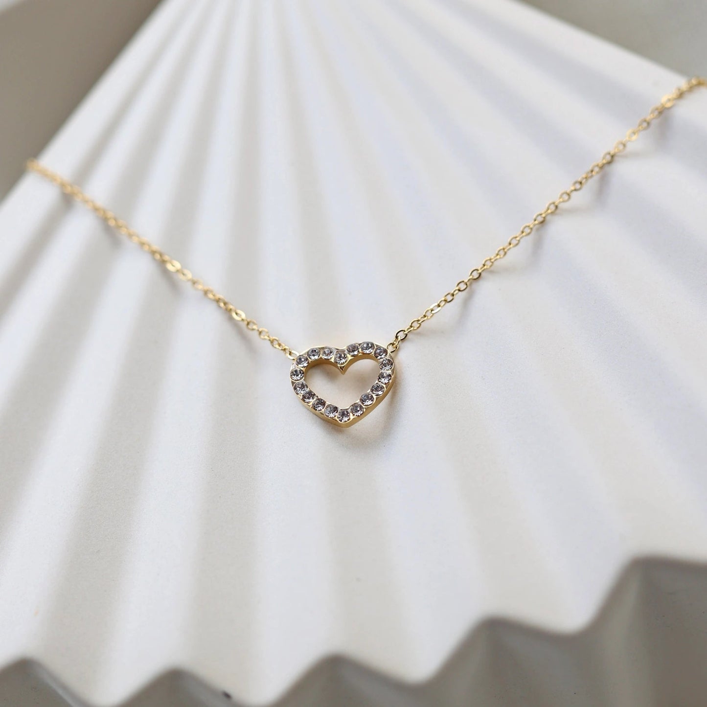 Collier Sentiment, pendentif cœur serti de brillants en acier inoxydable doré, photographié en gros plan sur un support beige texturé.