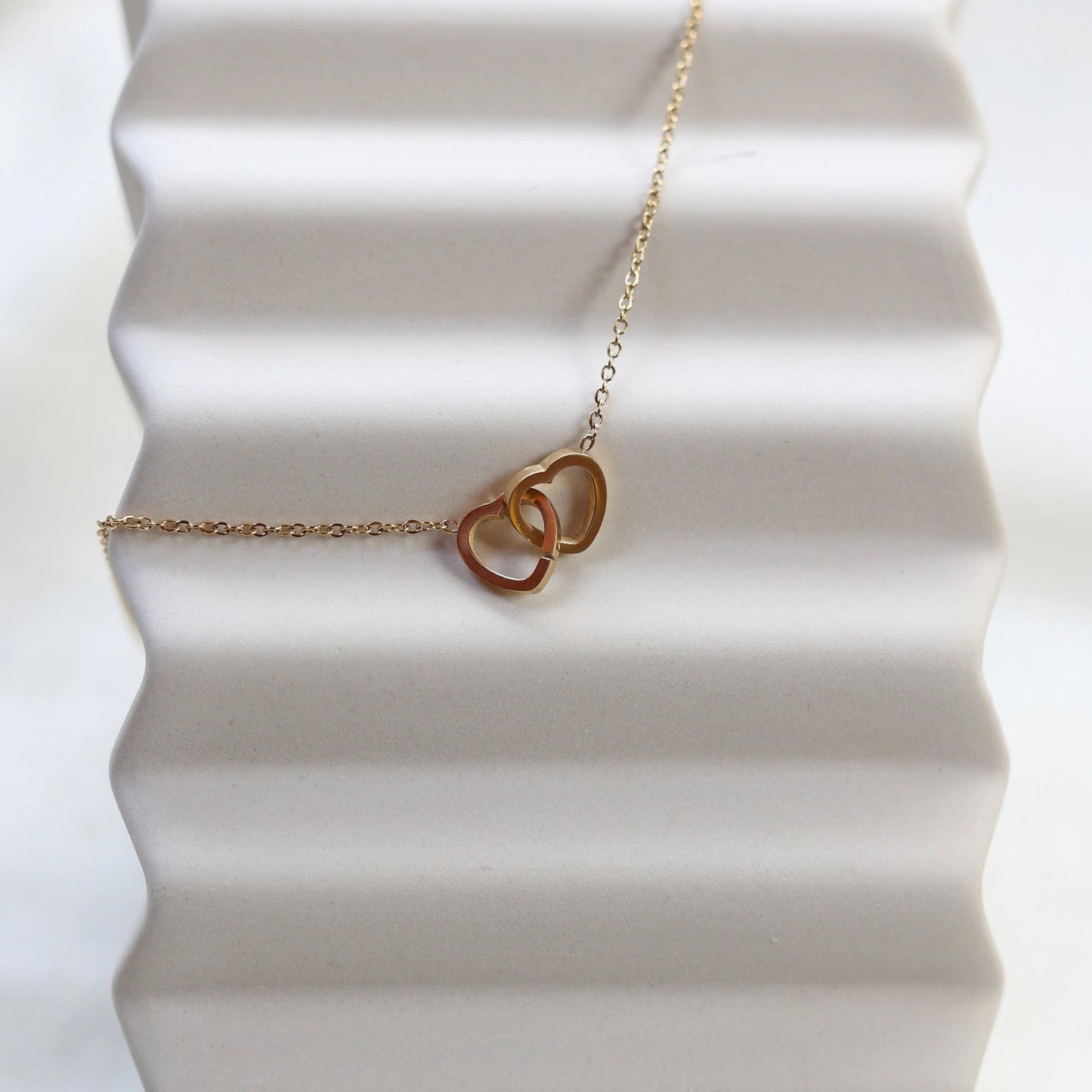 Collier Toi et moi en acier inoxydable doré, deux coeurs entrelacés, présenté en gros plan sur fond beige texturé.