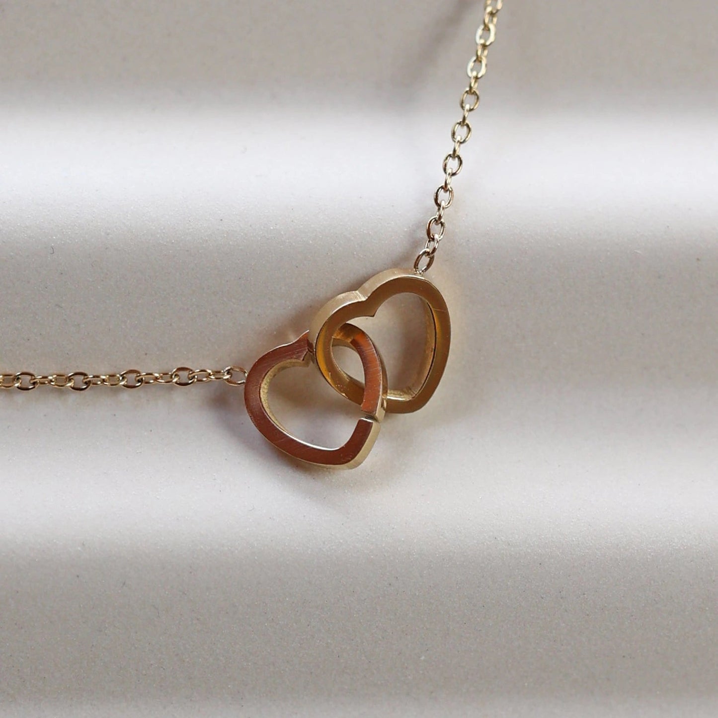 Collier Toi et moi en acier inoxydable doré, deux coeurs entrelacés, présenté en gros plan sur fond beige texturé.
