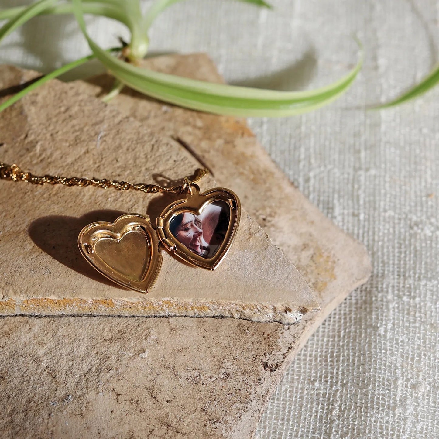 Zoom sur le collier "Entre nous", médaillon cœur en acier doré avec photo souvenir à personnaliser avec photo souvenir posé sur un fond beige texturé.