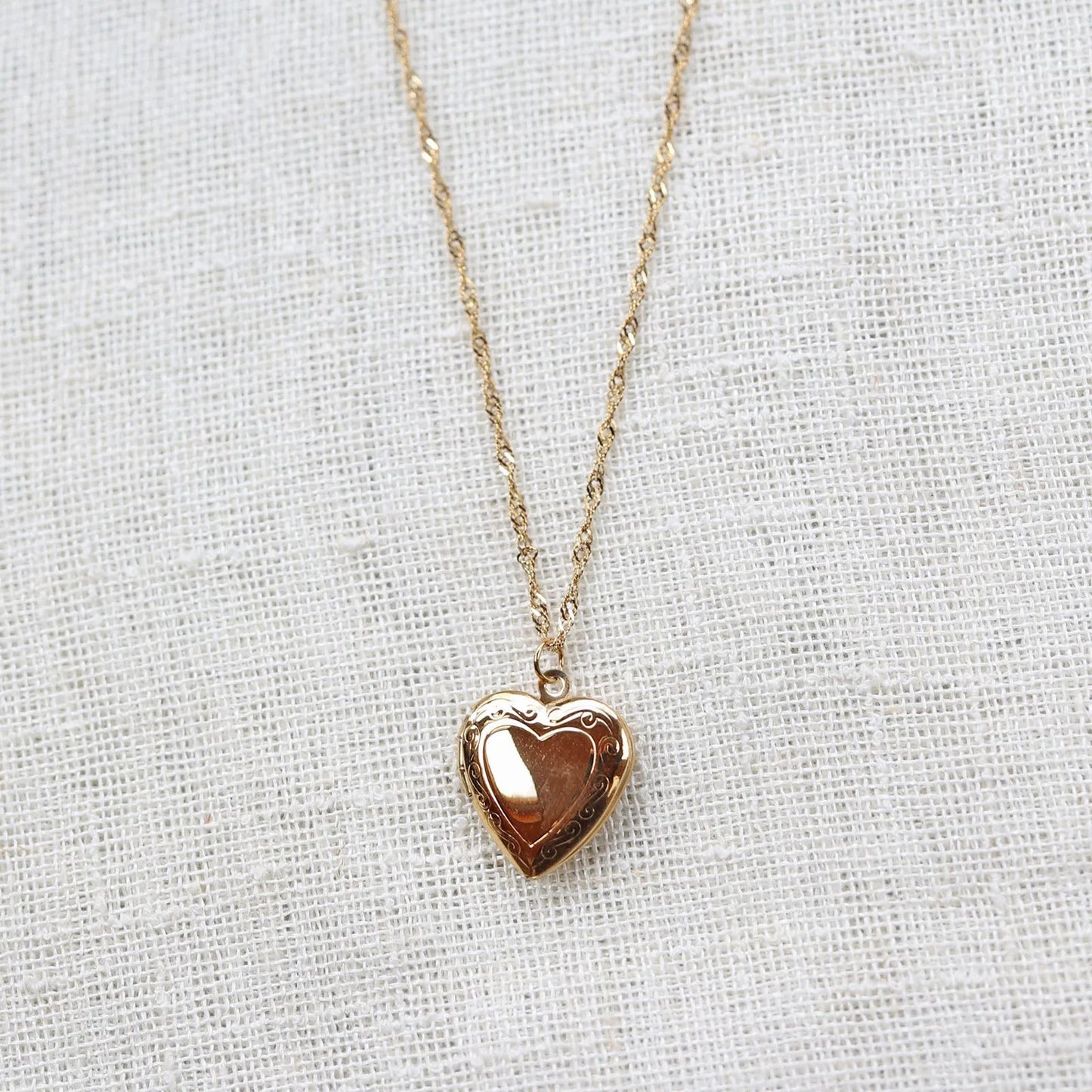 Collier "Entre nous", médaillon cœur en acier doré avec photo souvenir à personnaliser, posé sur un fond beige texturé.