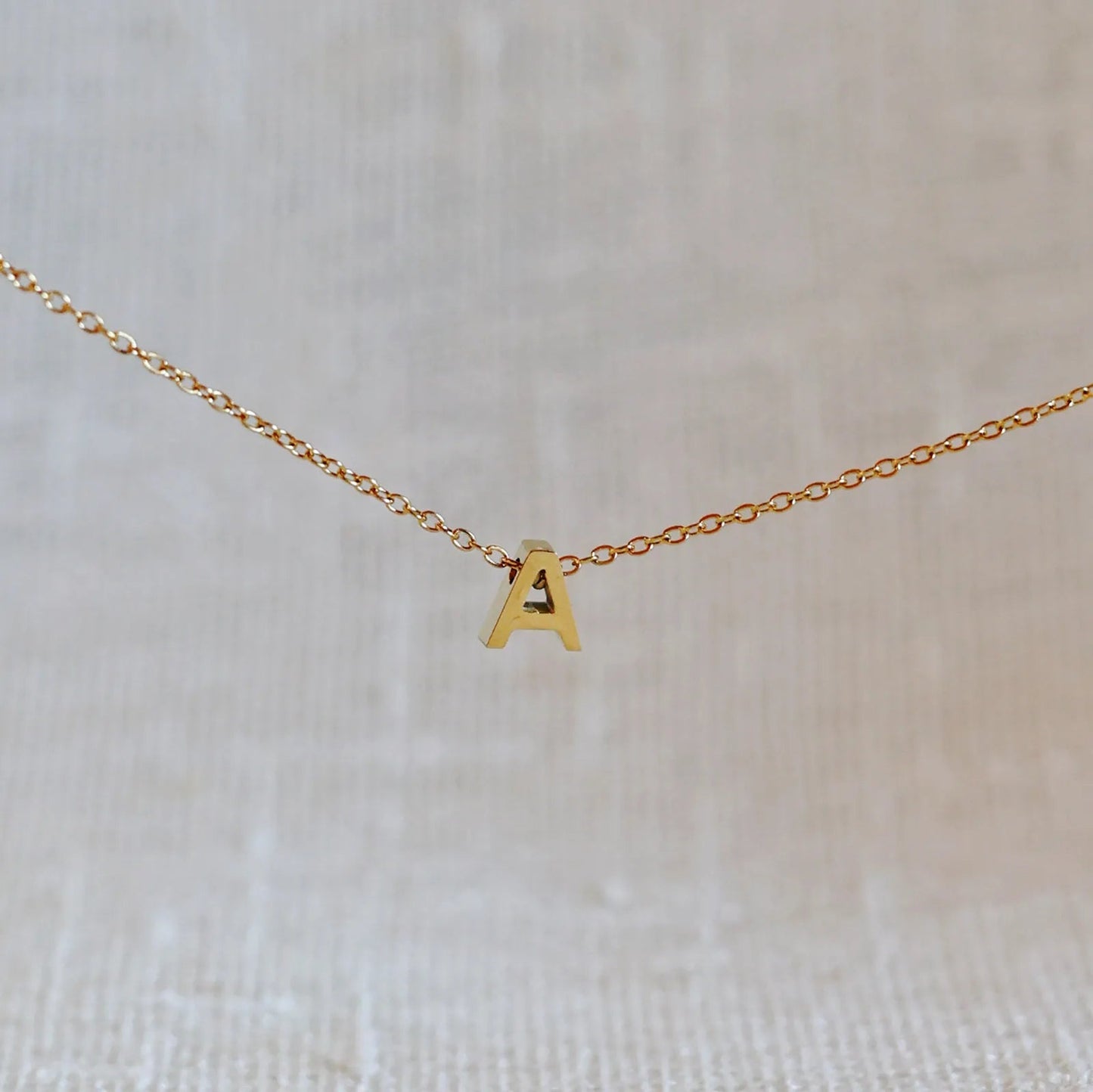Collier L'instant avec pendentif lettre A en acier inoxydable doré, bijou minimaliste suspendu sur fond beige.