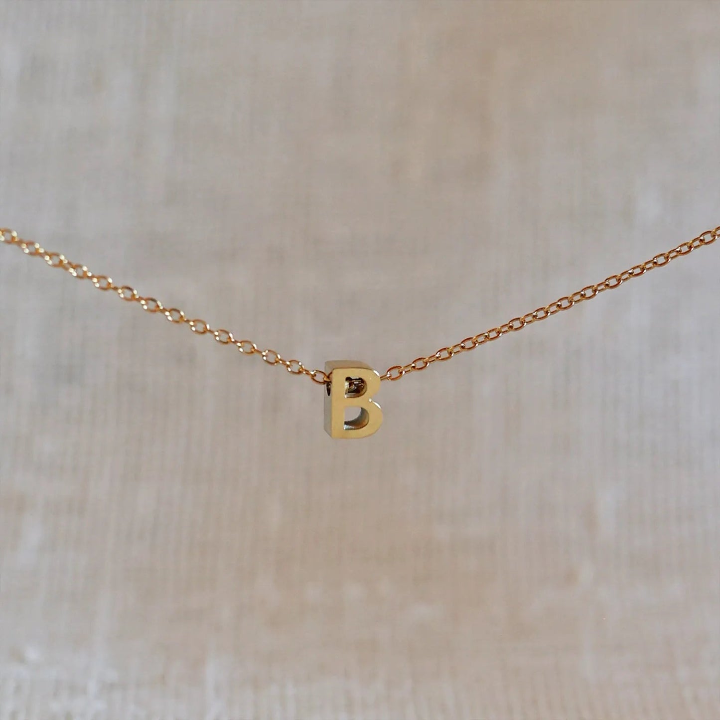 Collier L'instant avec pendentif lettre B doré en acier inoxydable, suspendu au centre d'une maille fine sur fond beige.