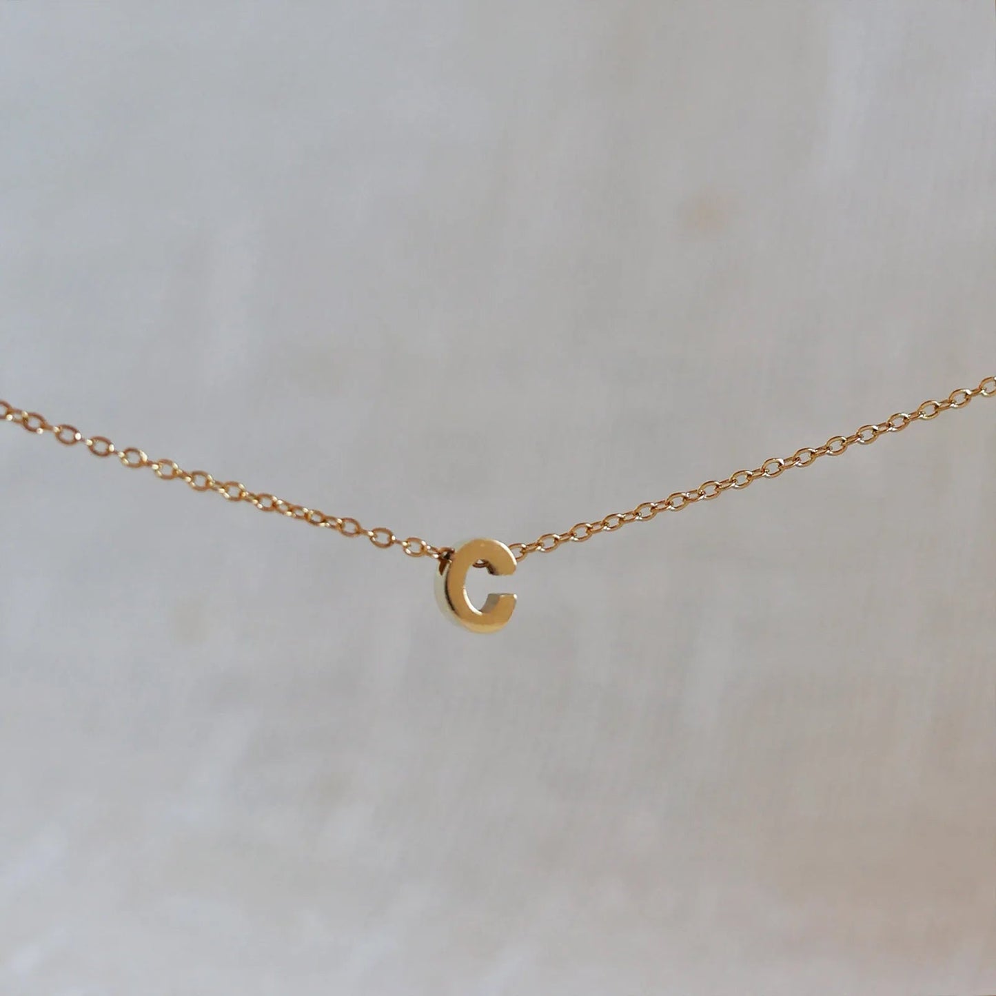 Collier L'instant avec pendentif lettre C doré en acier inoxydable, suspendu au centre d'une maille fine sur fond beige.
