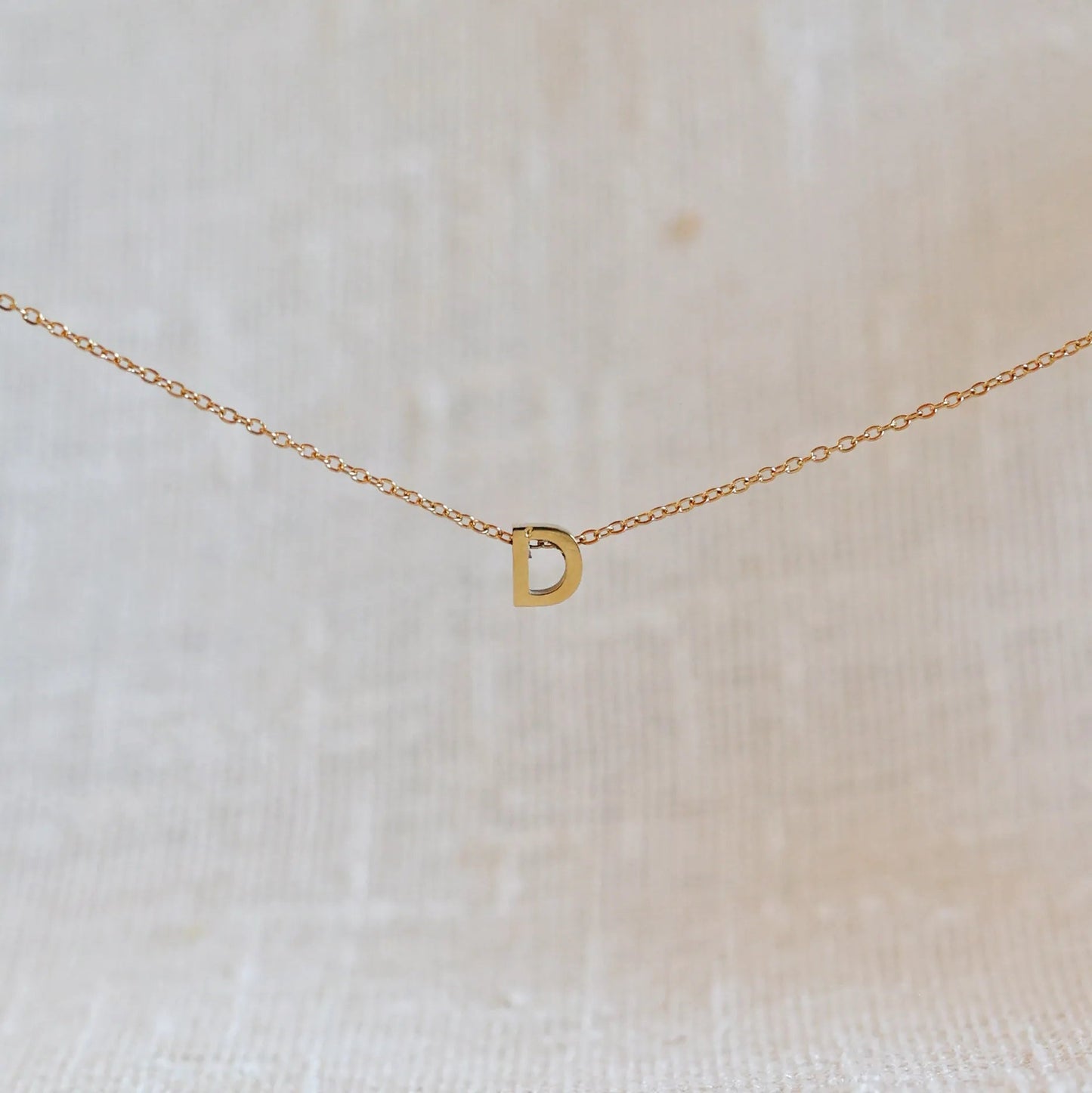 Collier L'instant avec pendentif lettre D doré en acier inoxydable, suspendu au centre d'une maille fine sur fond beige.