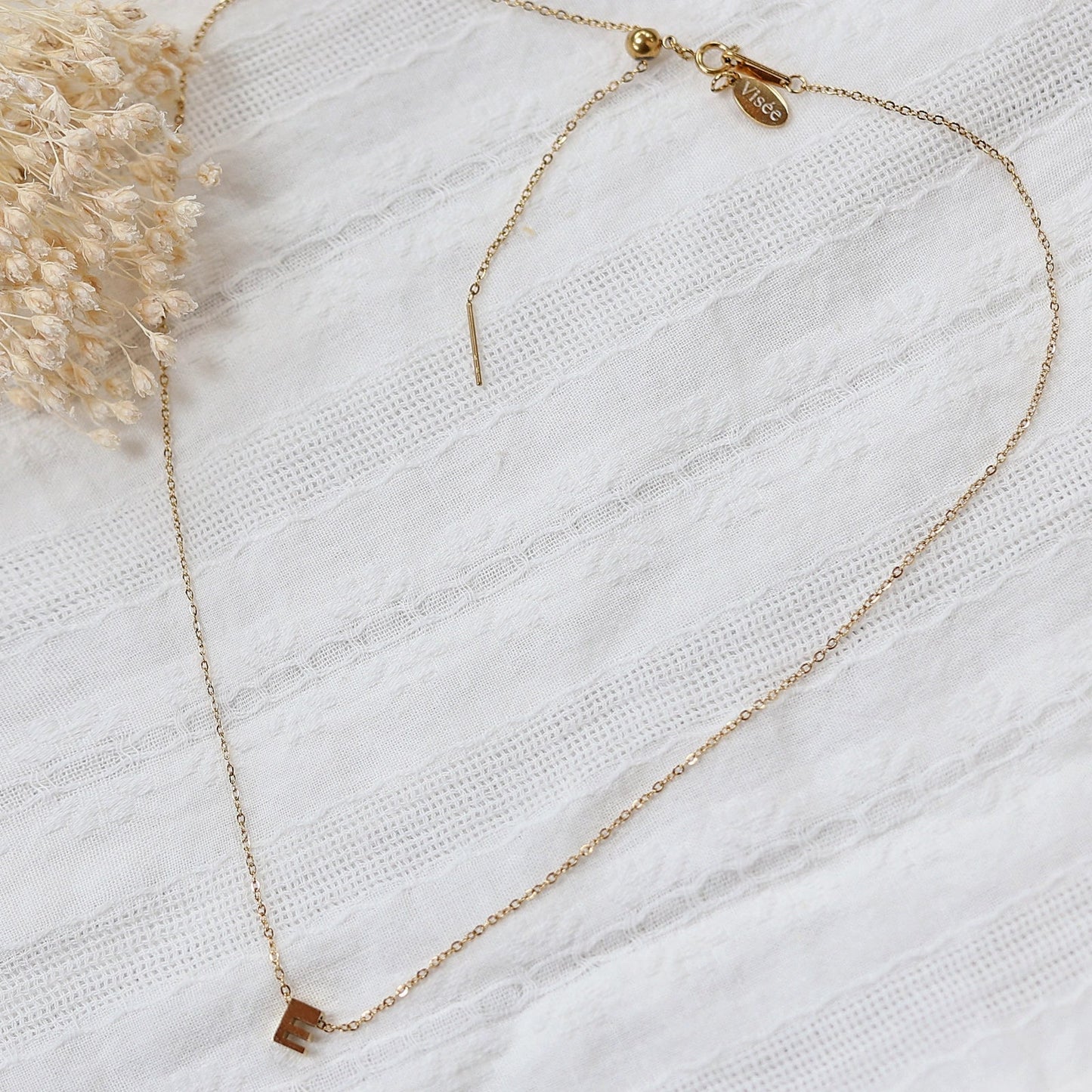 Collier L'instant avec pendentif lettre E en acier inoxydable doré, bijou inital délicat de Visée, photographié à plat sur un tissu beige doux.