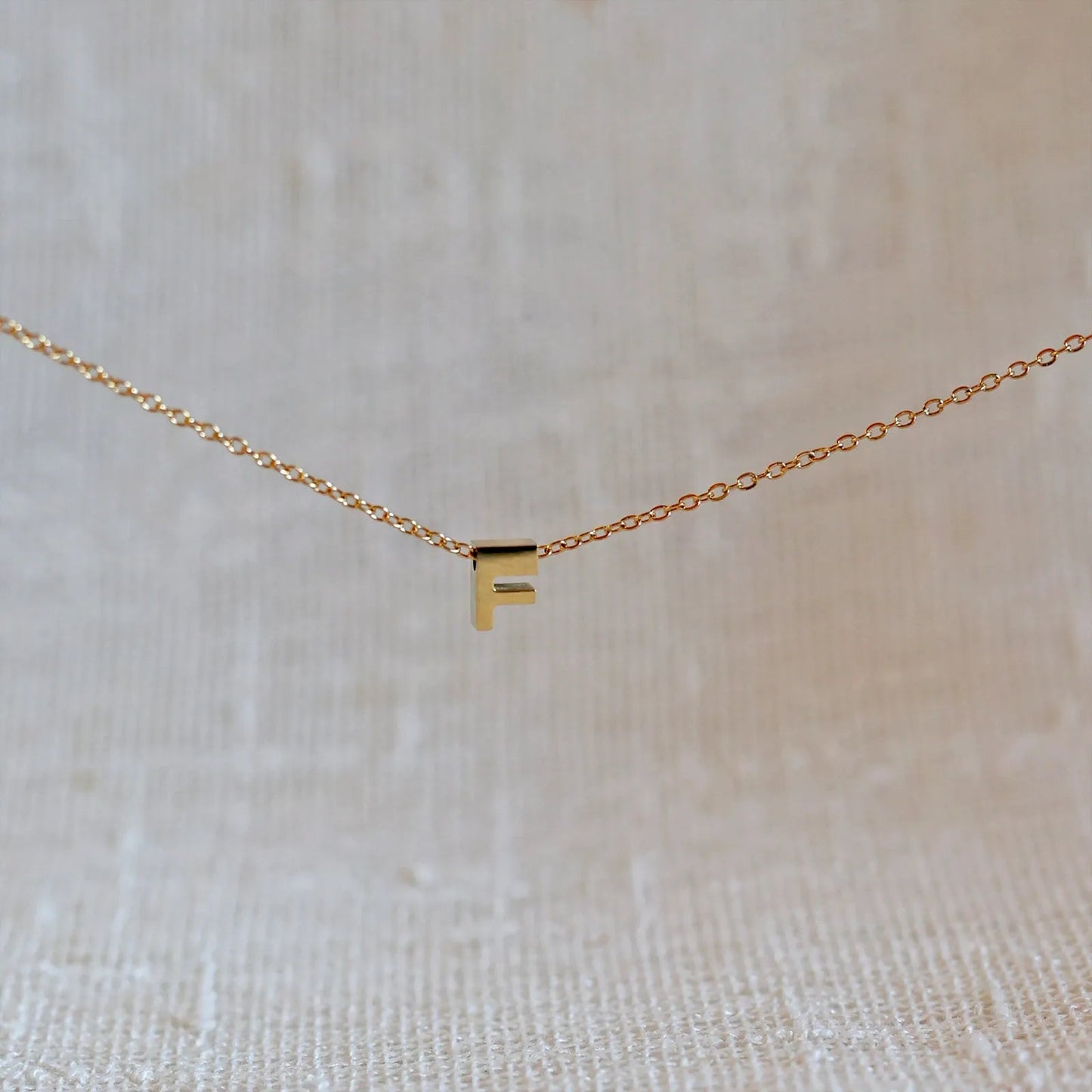 Collier L'instant avec pendentif lettre F doré en acier inoxydable, suspendu au centre d'une maille fine sur fond beige.