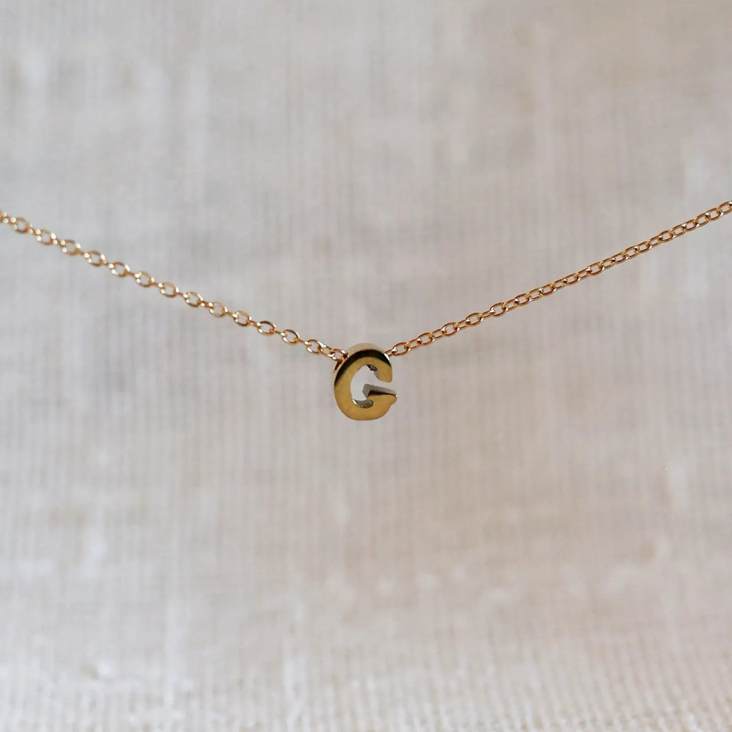 Collier L'instant avec pendentif lettre G doré en acier inoxydable, suspendu au centre d'une maille fine sur fond beige.